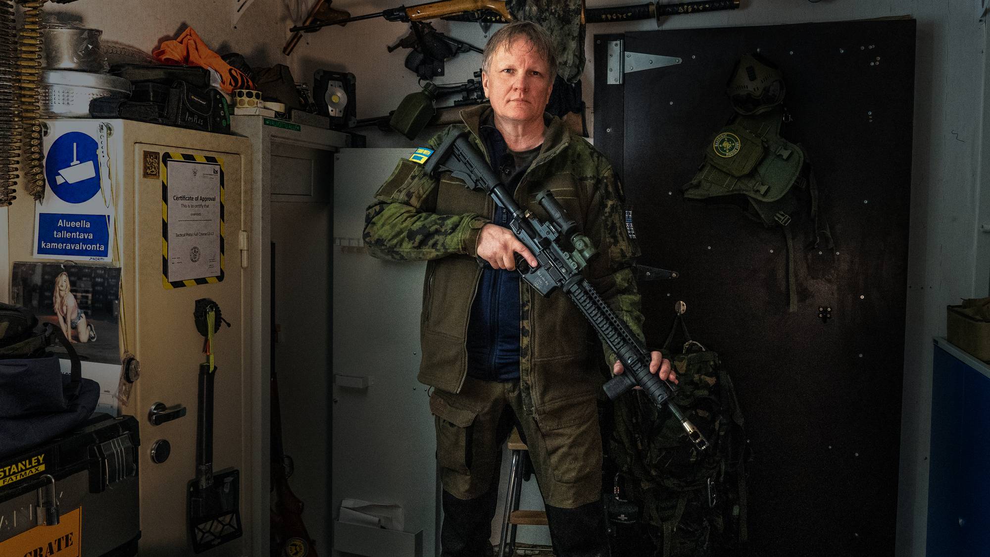 En person i militæruniform holder et automatgevær foran en dør. Rommet inneholder flere våpen, en safe og militært utstyr. Bildet er tatt i et mørkt og trangt rom. (Bildebeskrivelsen er laget av en KI-tjeneste)