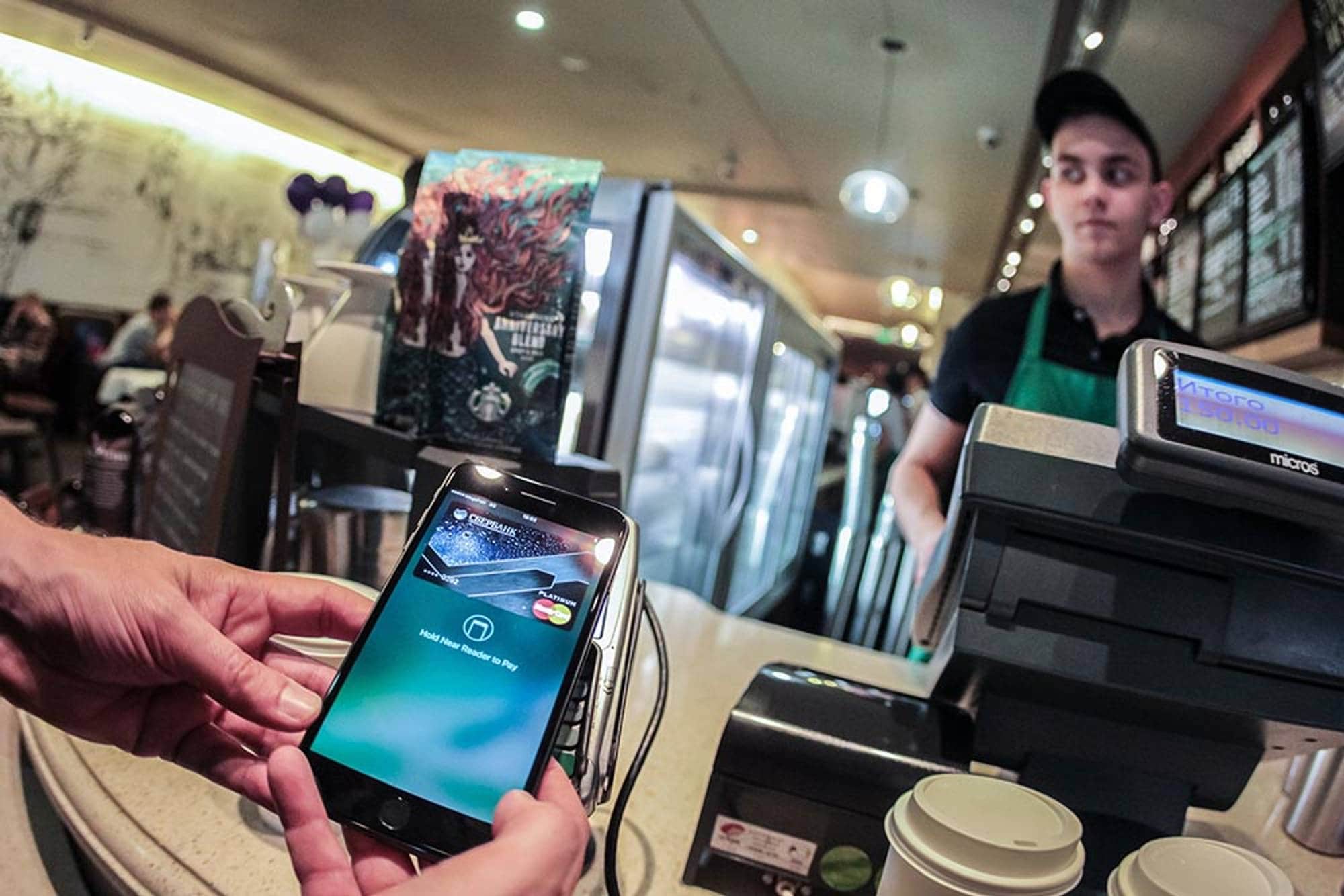 En kunde holder en mobiltelefon opp mot en betalingsterminal for å betale. En ansatt står bak disken på en kafé. Bakgrunnen viser andre gjester og kaféinnredning. (Bildebeskrivelsen er laget av en KI-tjeneste)