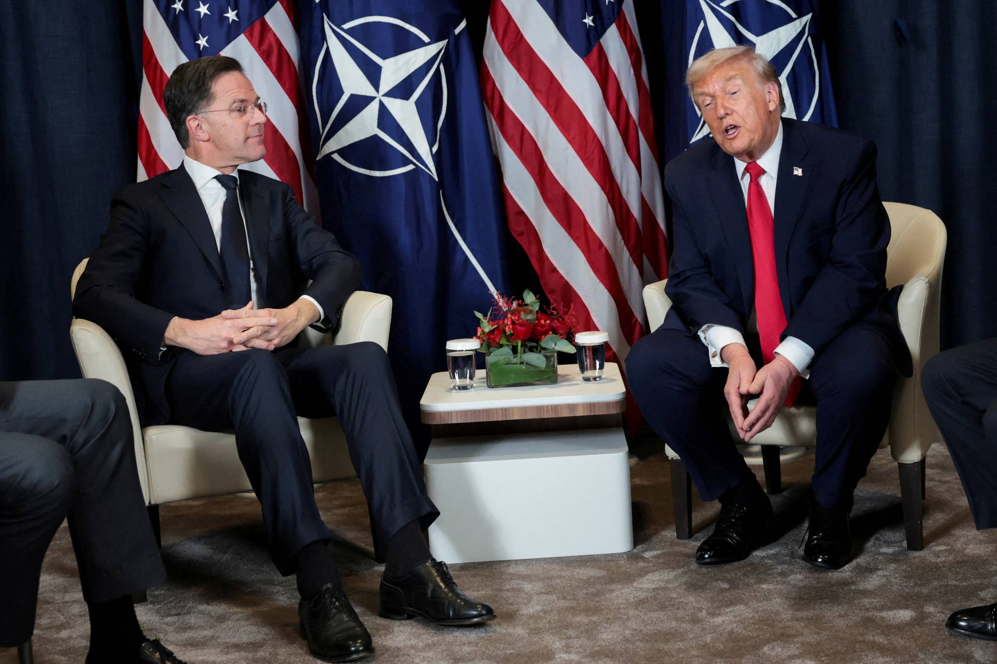 Nederlands statsminister Mark Rutte sitter sammen med USAs president Donald Trump. De sitter foran amerikanske og NATO-flagg. (Bildebeskrivelsen er laget av en KI-tjeneste)