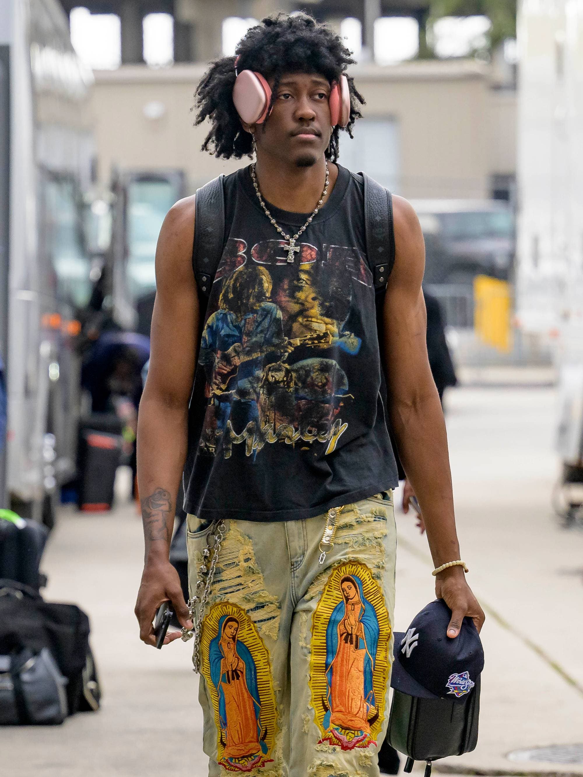 En mann med store hodetelefoner går forbi busser. Han har på seg en singlet med trykk og buksene har bilder av Jomfru Maria. En annen mann står med bagasje ved en av bussene. (Bildebeskrivelsen er laget av en KI-tjeneste)