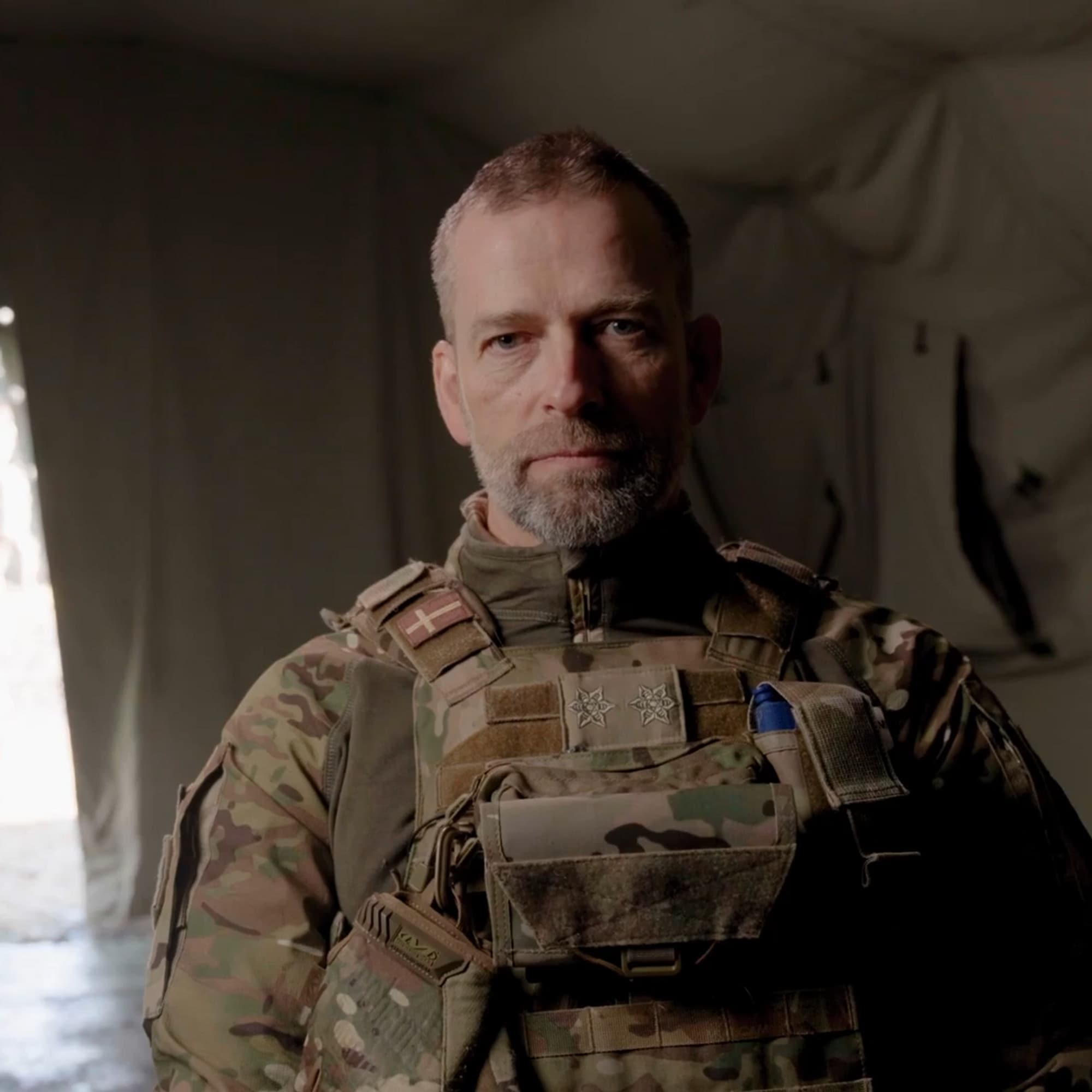 Generalmajoren er kledd i militæruniform og sitter i et telt med militærutstyr.