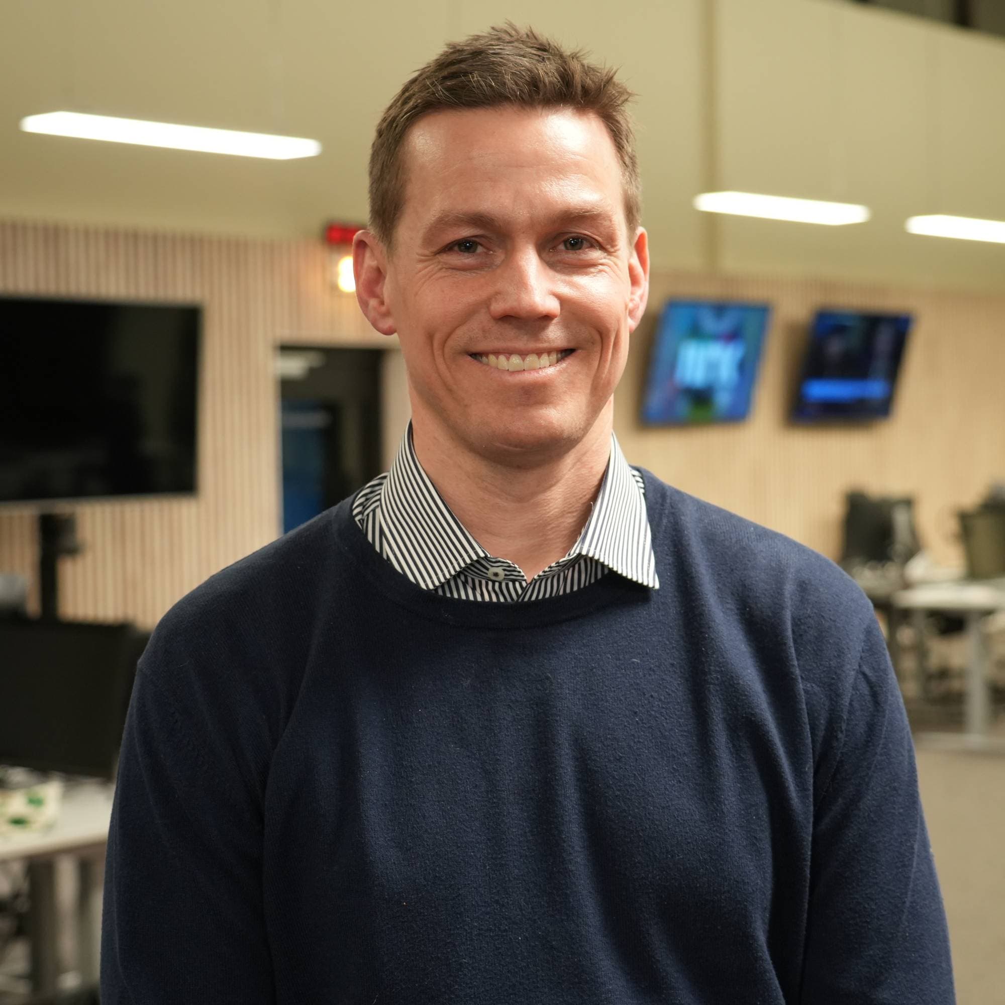 En mann med kort, brunt hår og blå genser smiler. Bak ham er et kontorlandskap med flere skjermer. (Bildebeskrivelsen er laget av en KI-tjeneste)