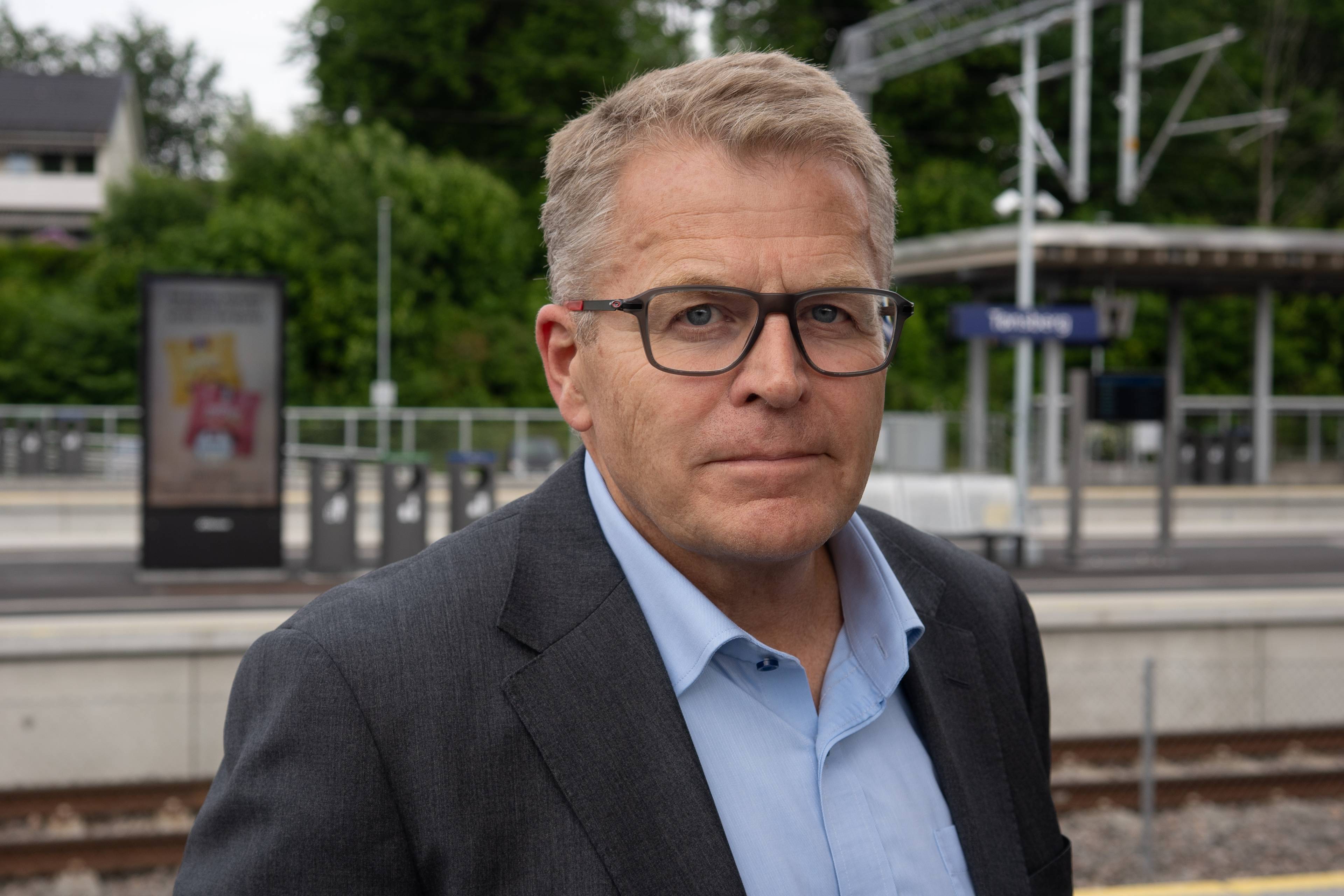 Mann med briller og mørk dressjakke står på en togstasjon. I bakgrunnen ses et perrongtak med et skilt hvor det står "Tønsberg".