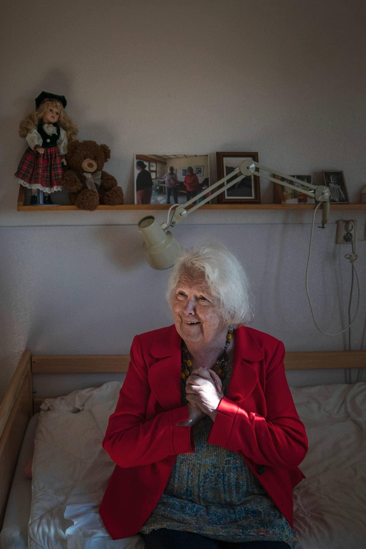 Kvinne i rød jakke sitter i seng og knytter hendene mens hun smiler stort. På veggen bak ser man en hylle med en rekke fotografier på en, en dukke og en teddybjørn.