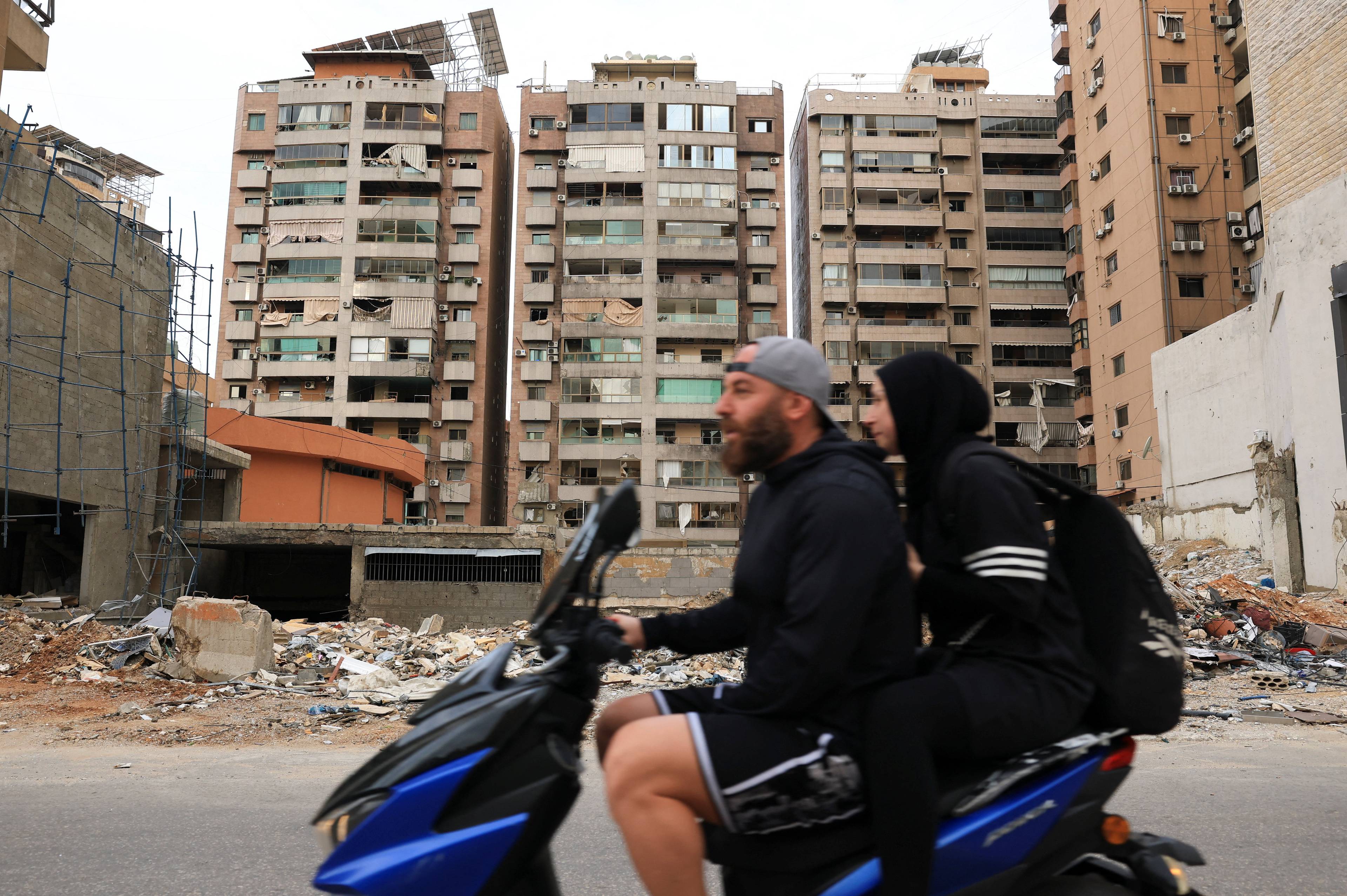 Et par kjører moped forbi bygninger med skader og rester etter ødeleggelse. En mann med caps sitter foran og en kvinne med hijab bak. Scener fra Gaza. (Bildebeskrivelsen er laget av en KI-tjeneste)