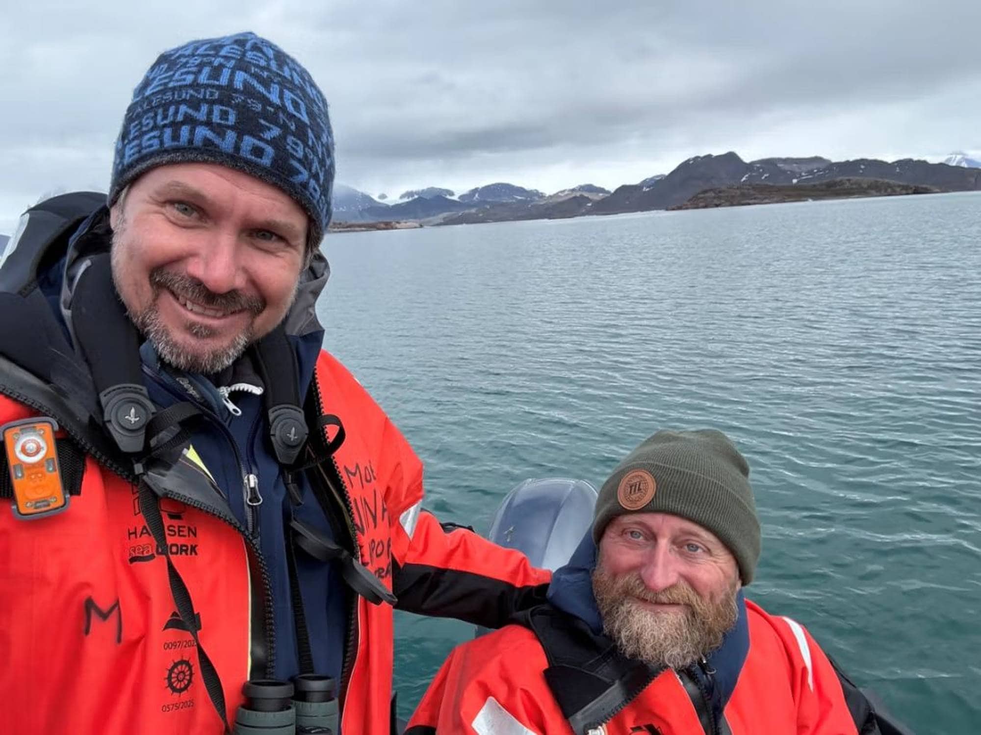 To menn i oransje flytedrakter i en båt på sjøen, med arktisk landskap i bakgrunnen. (Bildebeskrivelsen er laget av en KI-tjeneste)