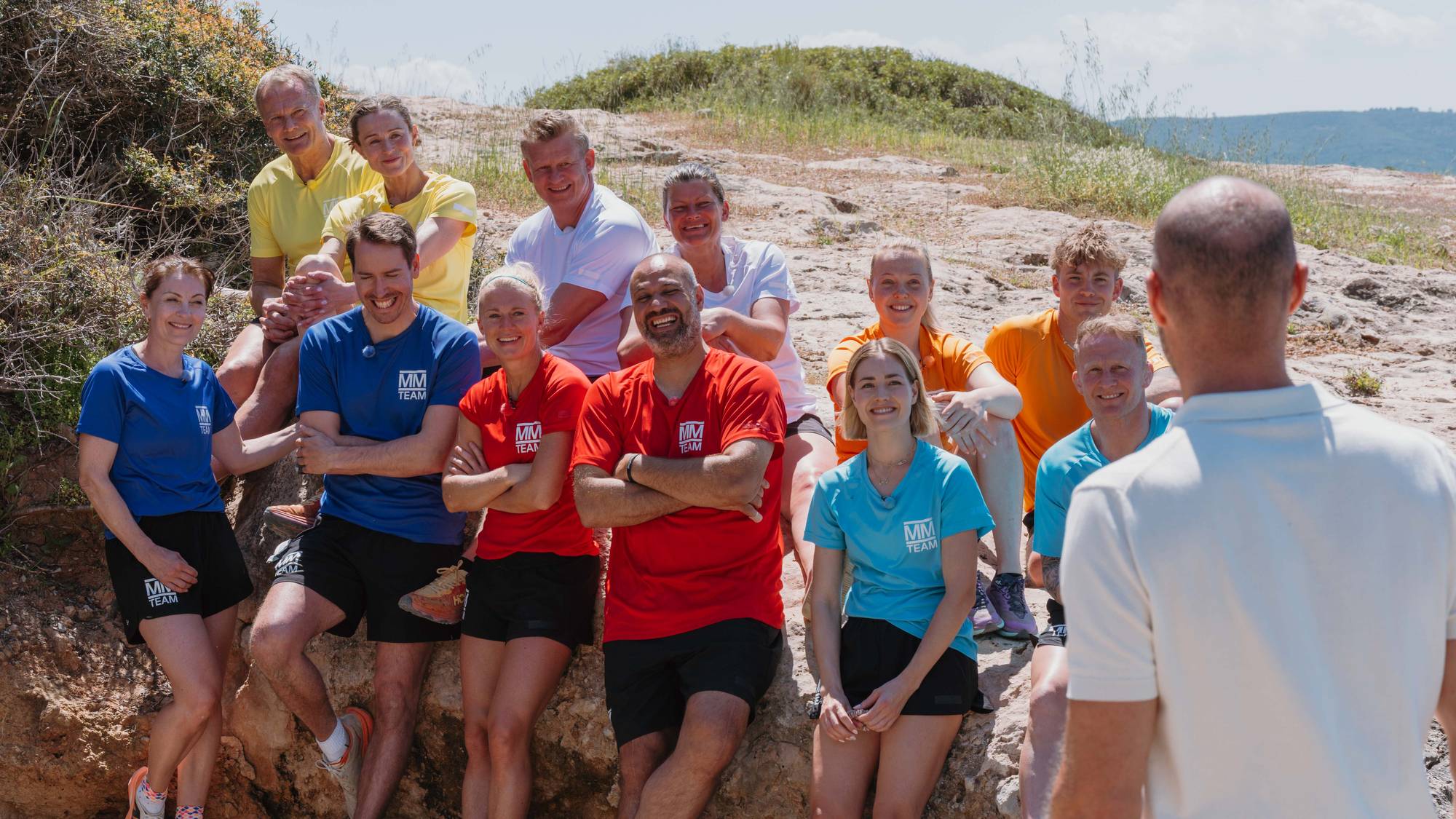 Tolv personer i fargede trøyer sittende på en knaus på stranden med sand og hav bak. Programleder i hvit trøye med ryggen til.