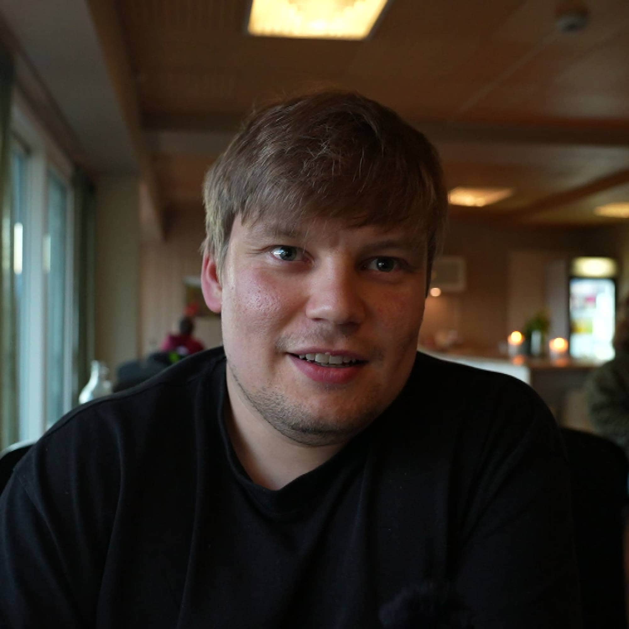 En mann som smiler til kameraet. I bakgrunnet er det et menneske som sitter ved et bord.