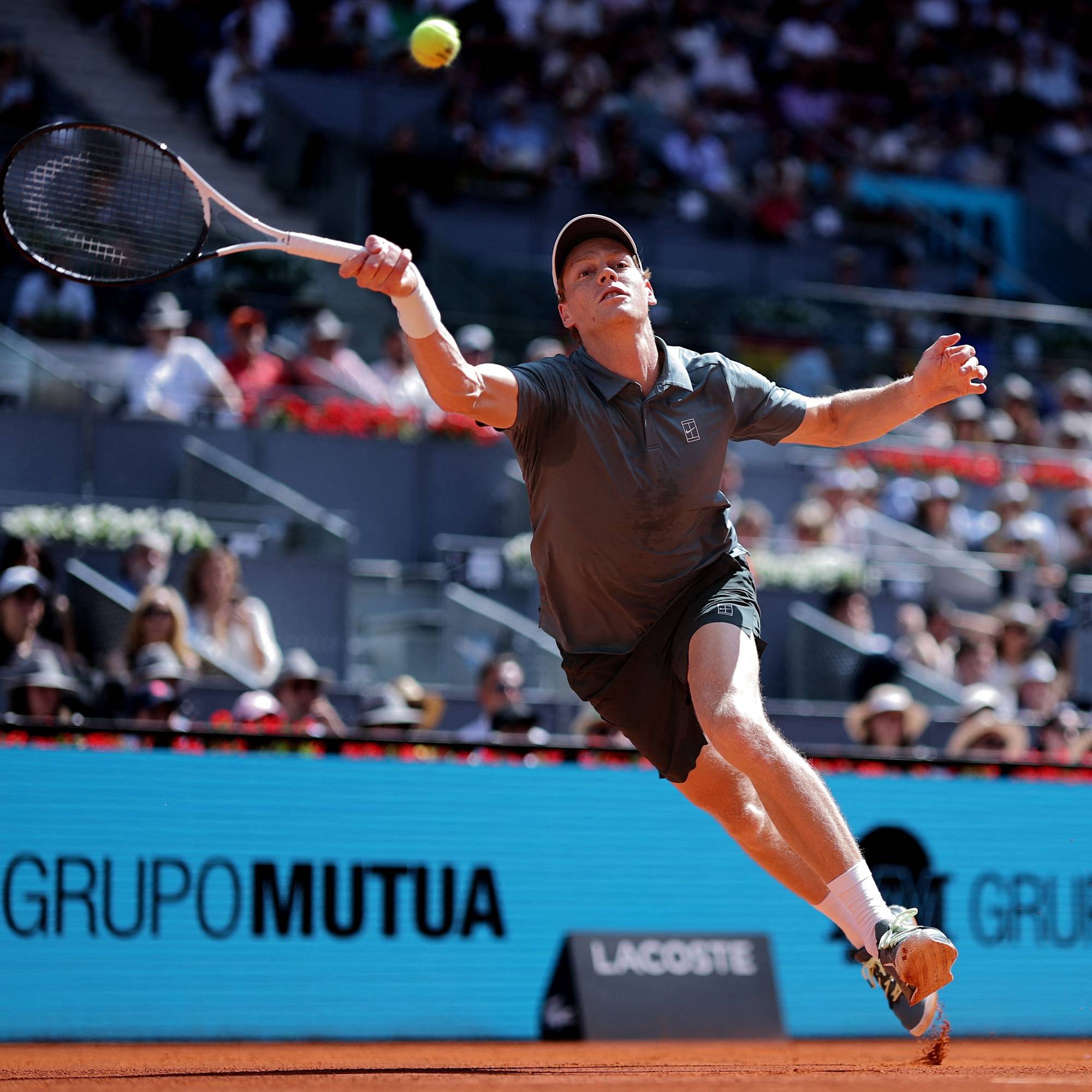 En mann spiller tennis på en rød grusbane. Han er iført en grå t-skjorte og mørke shorts, og har en racket i hånden. En tennisball flyr i luften over ham. Tilskuerne sitter på tribunene i bakgrunnen. (Bildebeskrivelsen er laget av en KI-tjeneste)