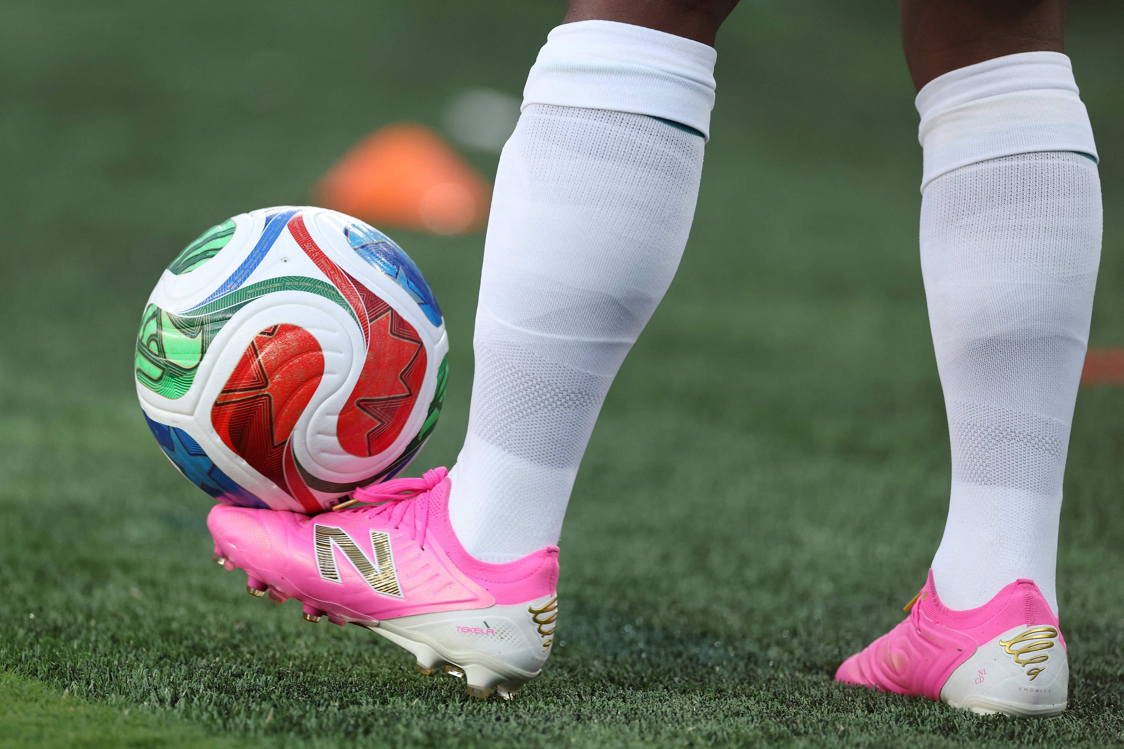 Nærbilde av en fotballspillers føtter. Spilleren har hvite sokker og rosa fotballsko. En fotball ligger på gresset ved skoen. I bakgrunnen er det en fotballbane med noen kjegler. (Bildebeskrivelsen er laget av en KI-tjeneste)