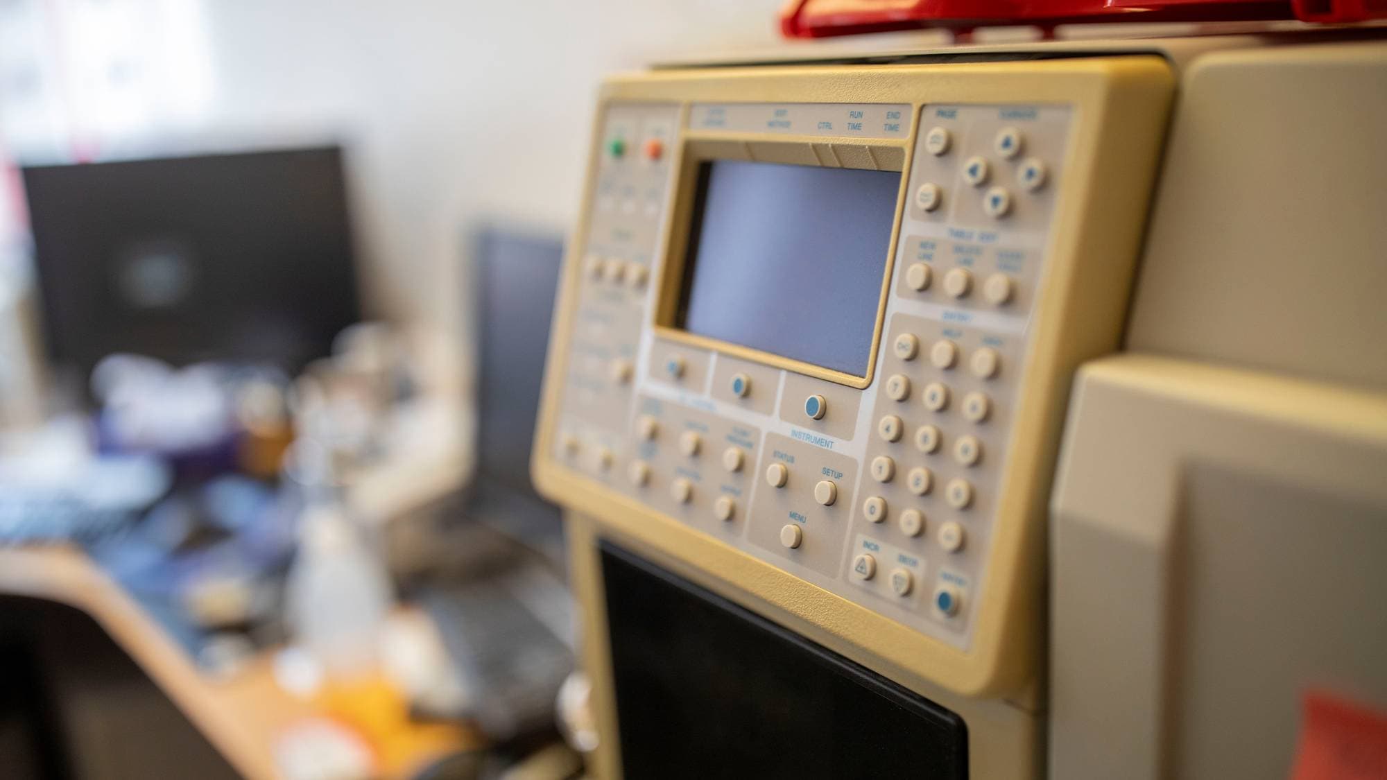 Et beige analyseinstrument med et digitalt display og mange knapper står i forgrunnen. Instrumentet er plassert på et kontor med dataskjermer og utstyr i bakgrunnen. Bildet er tatt tett på instrumentet. (Bildebeskrivelsen er laget av en KI-tjeneste)