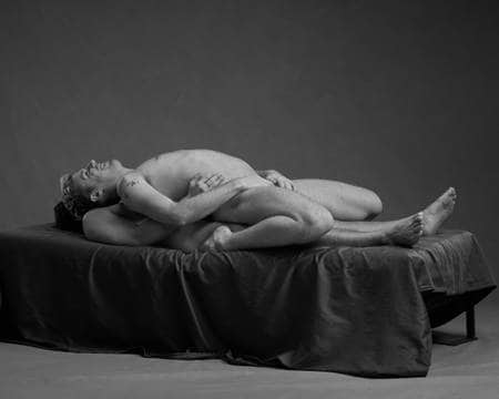 To nakne menn ligger på hverandre på en mørk seng. Den øverste mannen ligger med ryggen mot den andre, med beina bøyd og kroppen lent bakover. Han har hendene mellom beina og smiler. Den underste mannen har på siden av magen til den andre.