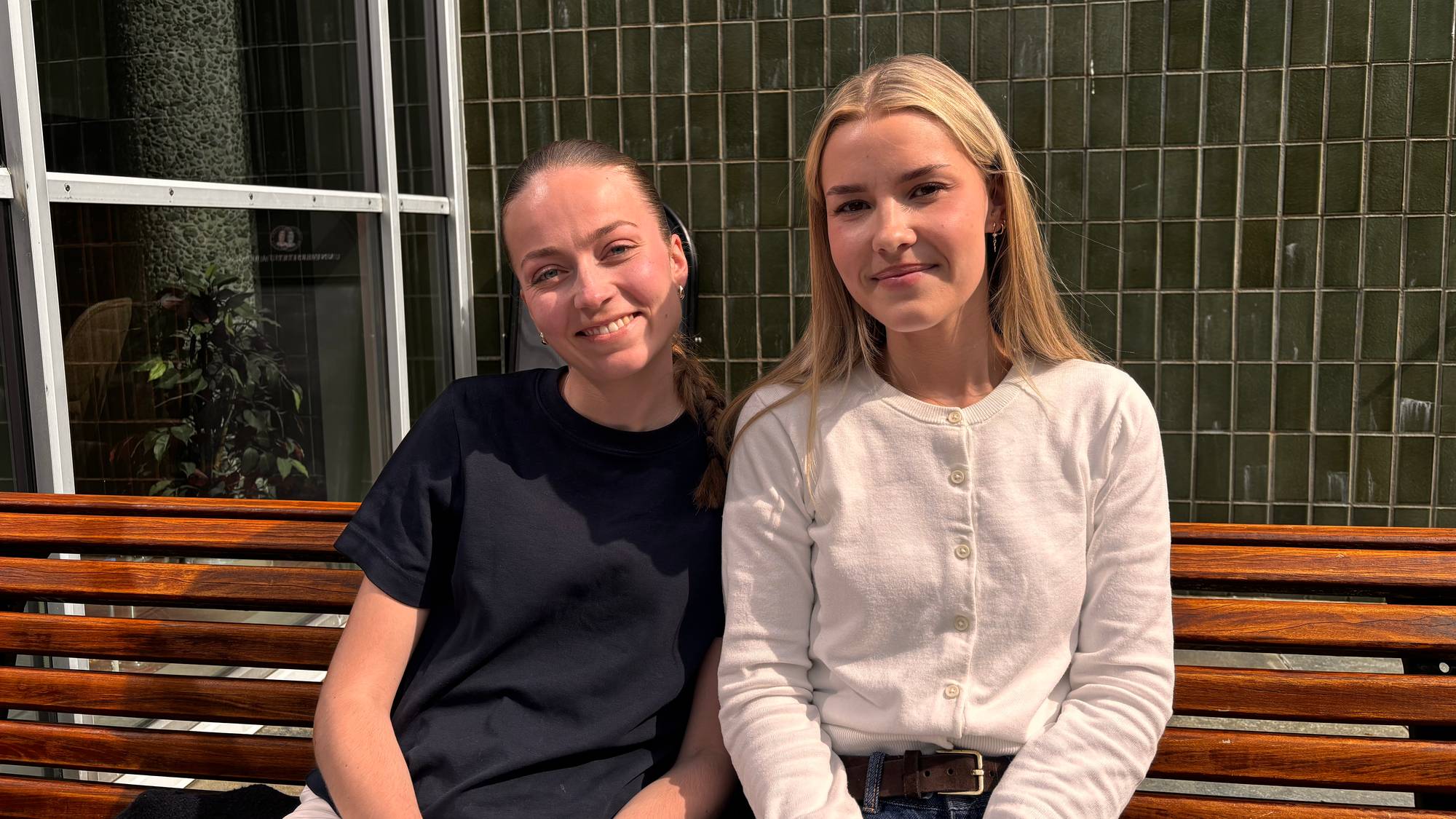 Two young women sit side-by-side on a wooden bench outdoors. The woman on the left wears a dark t-shirt and has her hair in a braid. The woman on the right wears a white cardigan. Behind them is a tiled wall. (Bildebeskrivelsen er laget av en KI-tjeneste)