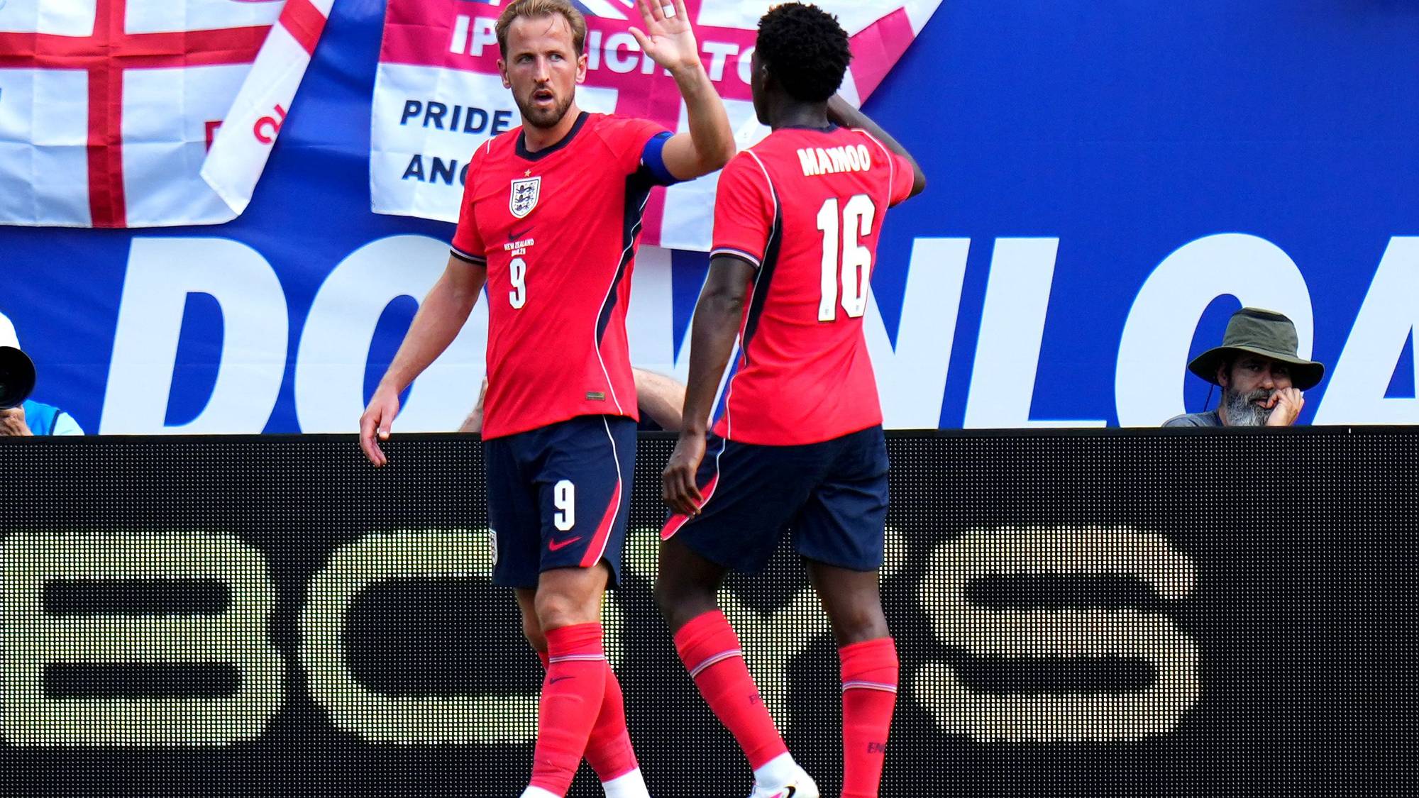 To fotballspillere, en med draktnummer 9 og en med 16, gir hverandre en high-five. Bak dem henger flere flagg, inkludert det britiske flagget. En mann med hatt sitter og ser ut tilskuere. (Bildebeskrivelsen er laget av en KI-tjeneste)