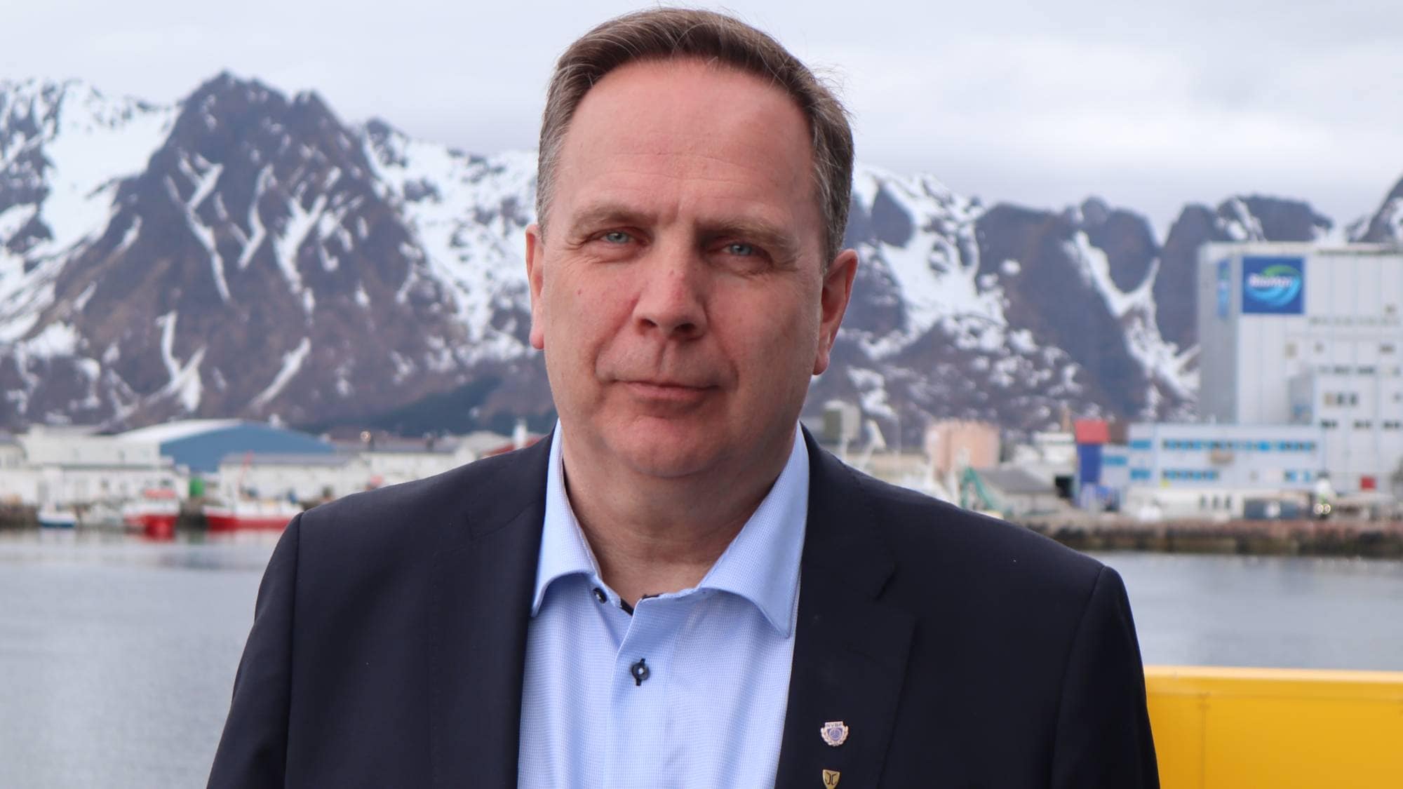 Mann i dress står foran en havn med snødekte fjell i bakgrunnen. (Bildebeskrivelsen er laget av en KI-tjeneste)
