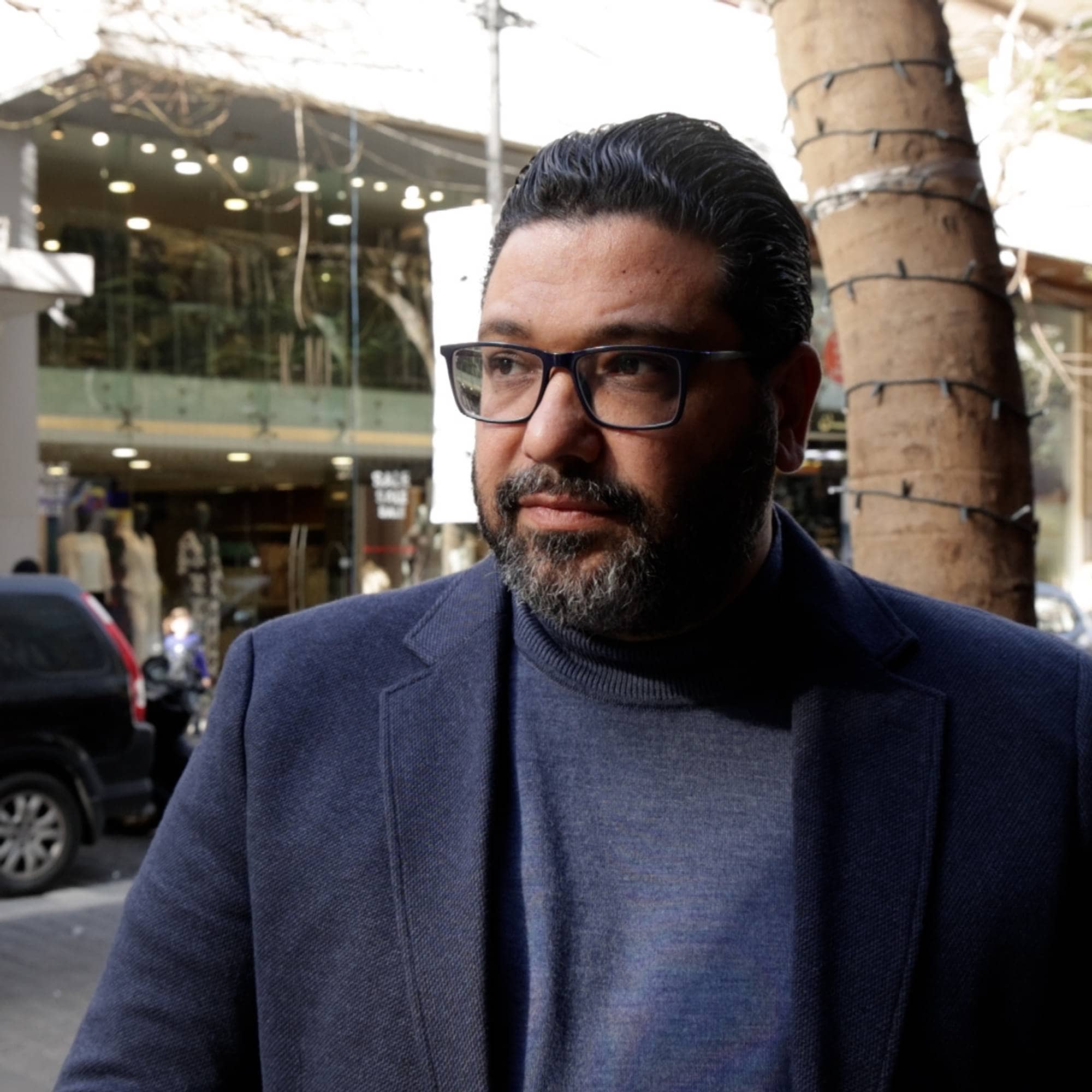 Mann med skjegg og briller i blå dressjakke, står i en bygate