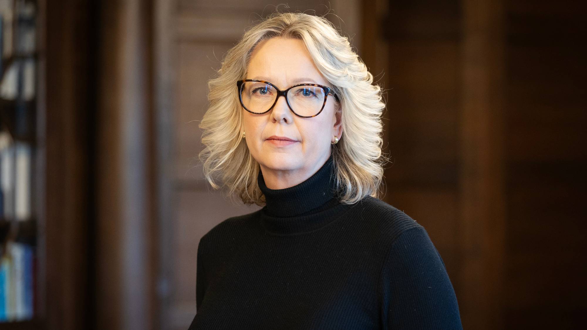 Portrett av statssekretær Eileen Fugelsnes med mørk brun bakgrunn. Hun har på seg en høyhalset sort genser og sorte briller. Håret er lyst og skulderlangt. 