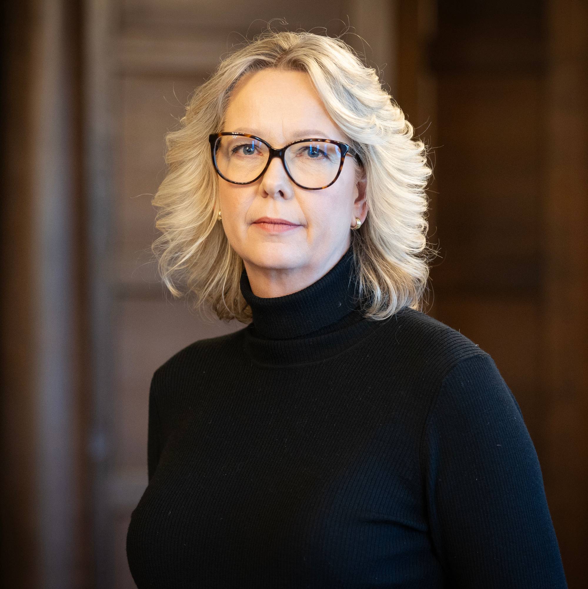 Portrett av statssekretær Eileen Fugelsnes med mørk brun bakgrunn. Hun har på seg en høyhalset sort genser og sorte briller. Håret er lyst og skulderlangt. 