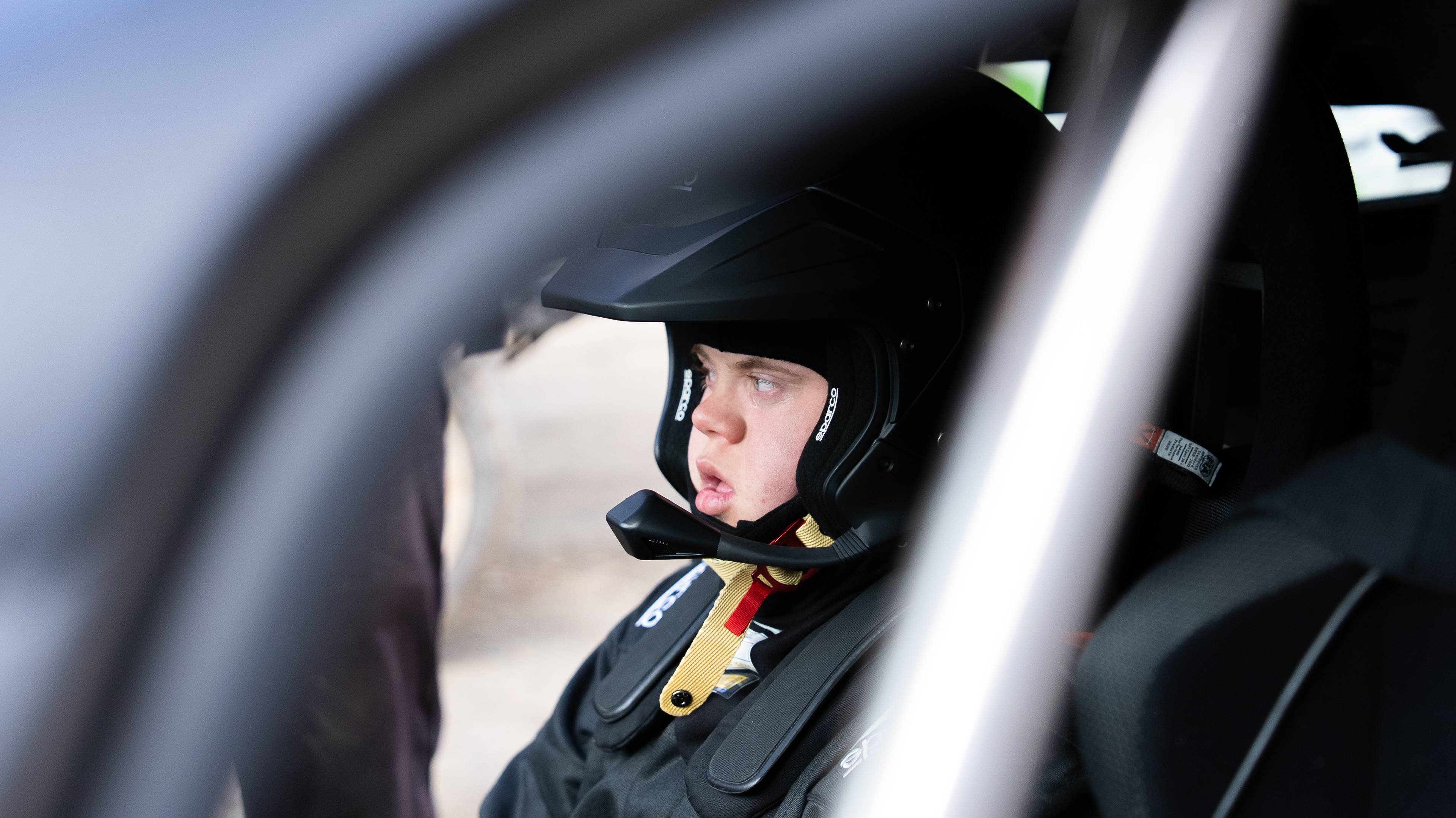 En person sitter i et racing-bil med hjelm og nakkebeskyttelse. Ansiktet er i profil. Rundt personen er bilens interiør synlig, med seter og bur. (Bildebeskrivelsen er laget av en KI-tjeneste)