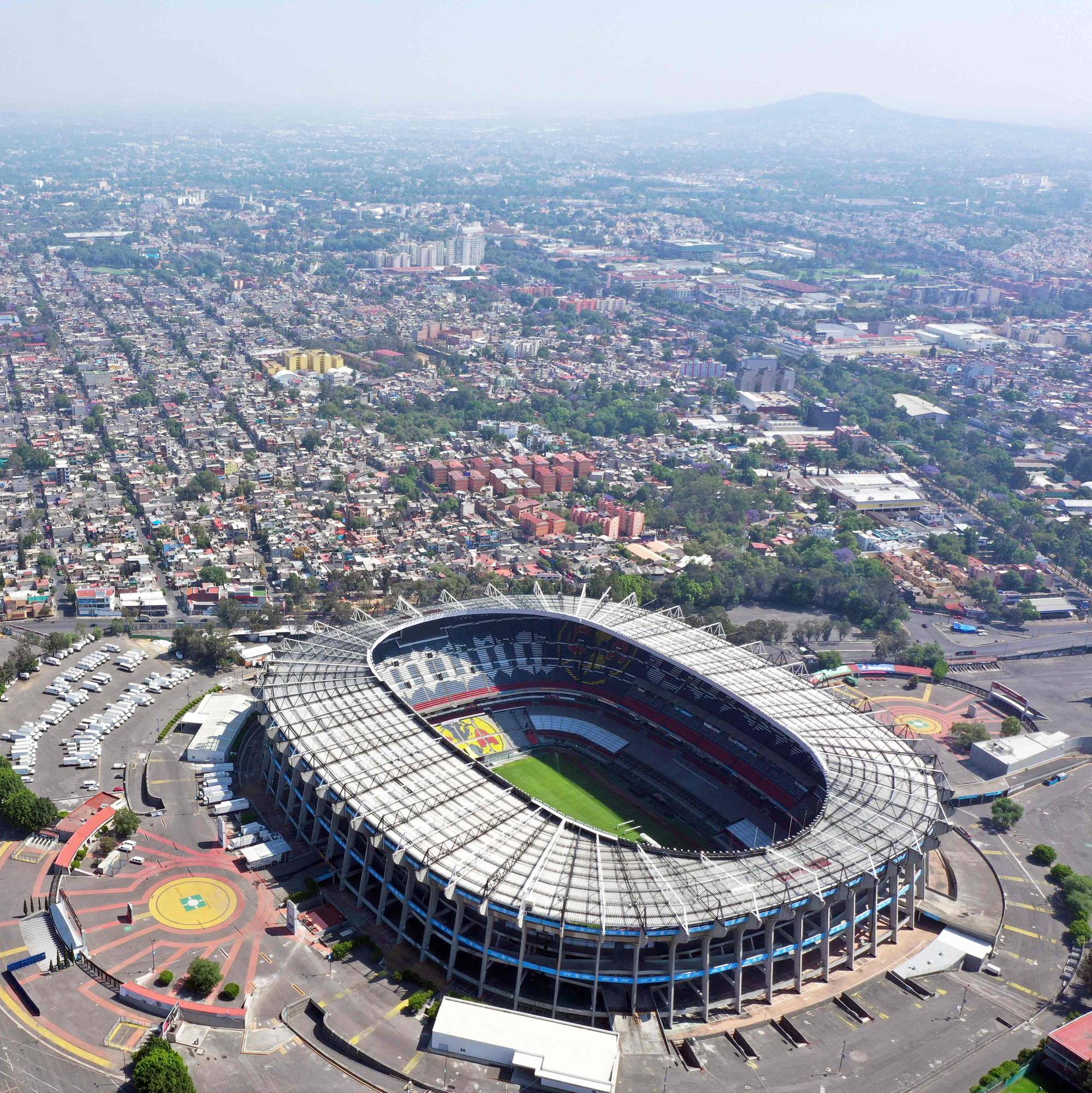 Dronefoto viser et stort stadion omkranset av en by. Stadionet har grønt gress og tribuner. Rundt stadionet er det en stor parkeringsplass og bygninger. (Bildebeskrivelsen er laget av en KI-tjeneste)