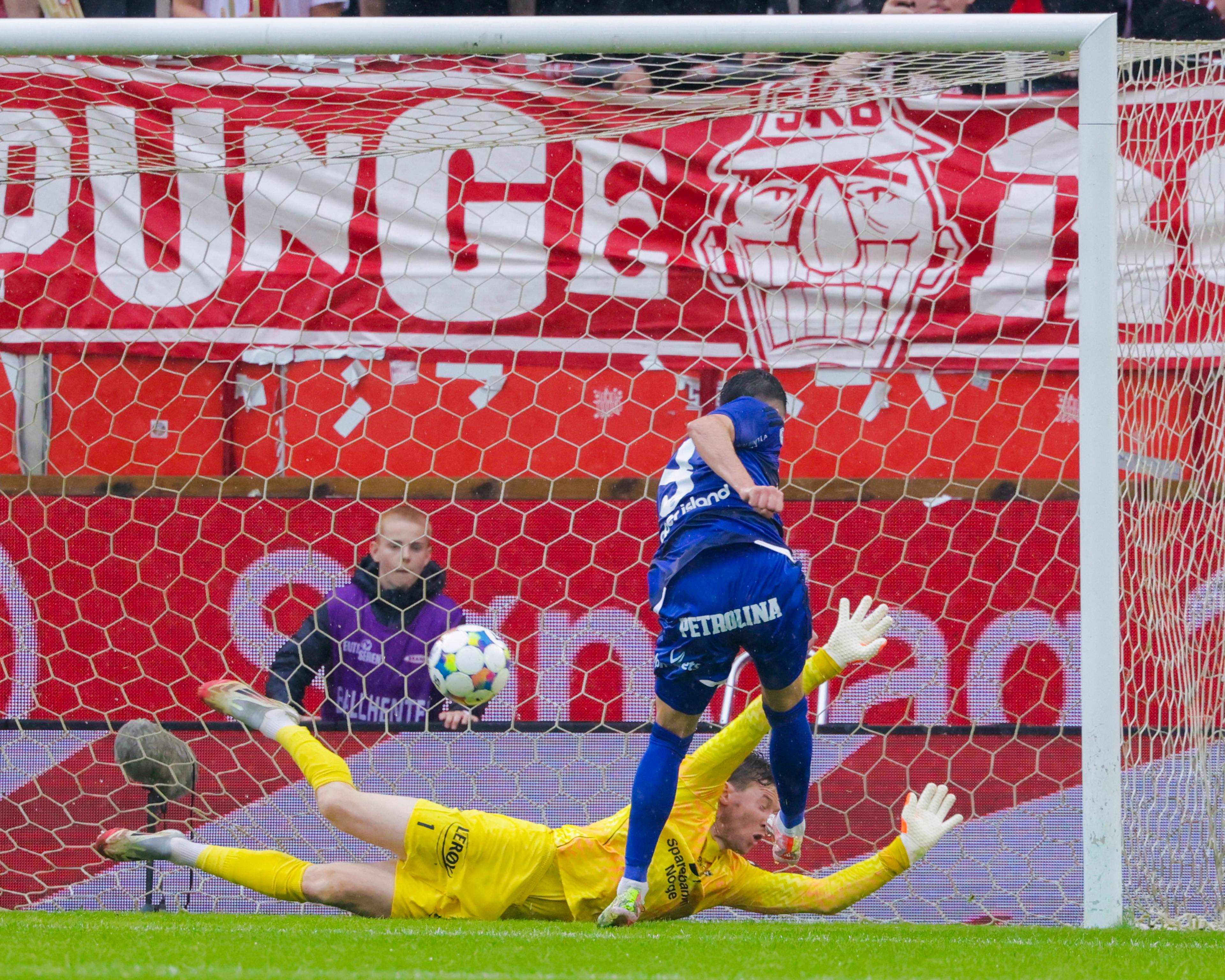 Fotballspiller i blått hopper over en keeper i gult under en kamp foran et rødt tribunebanner. (Bildebeskrivelsen er laget av en KI-tjeneste)