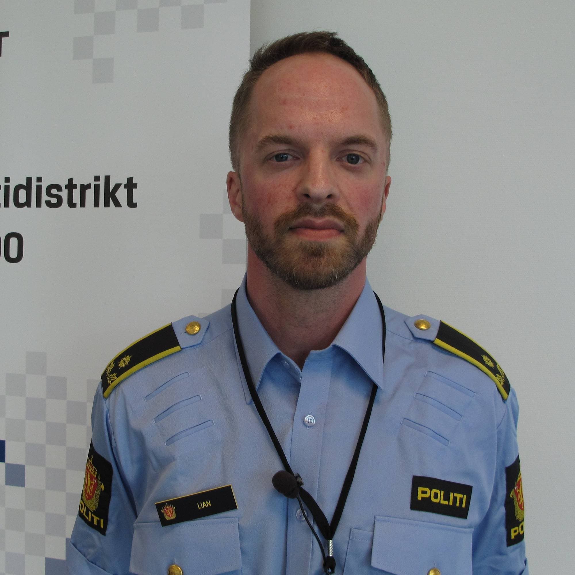 En mann med skjegg og politiuniform står foran en hvit vegg med politiinformasjon