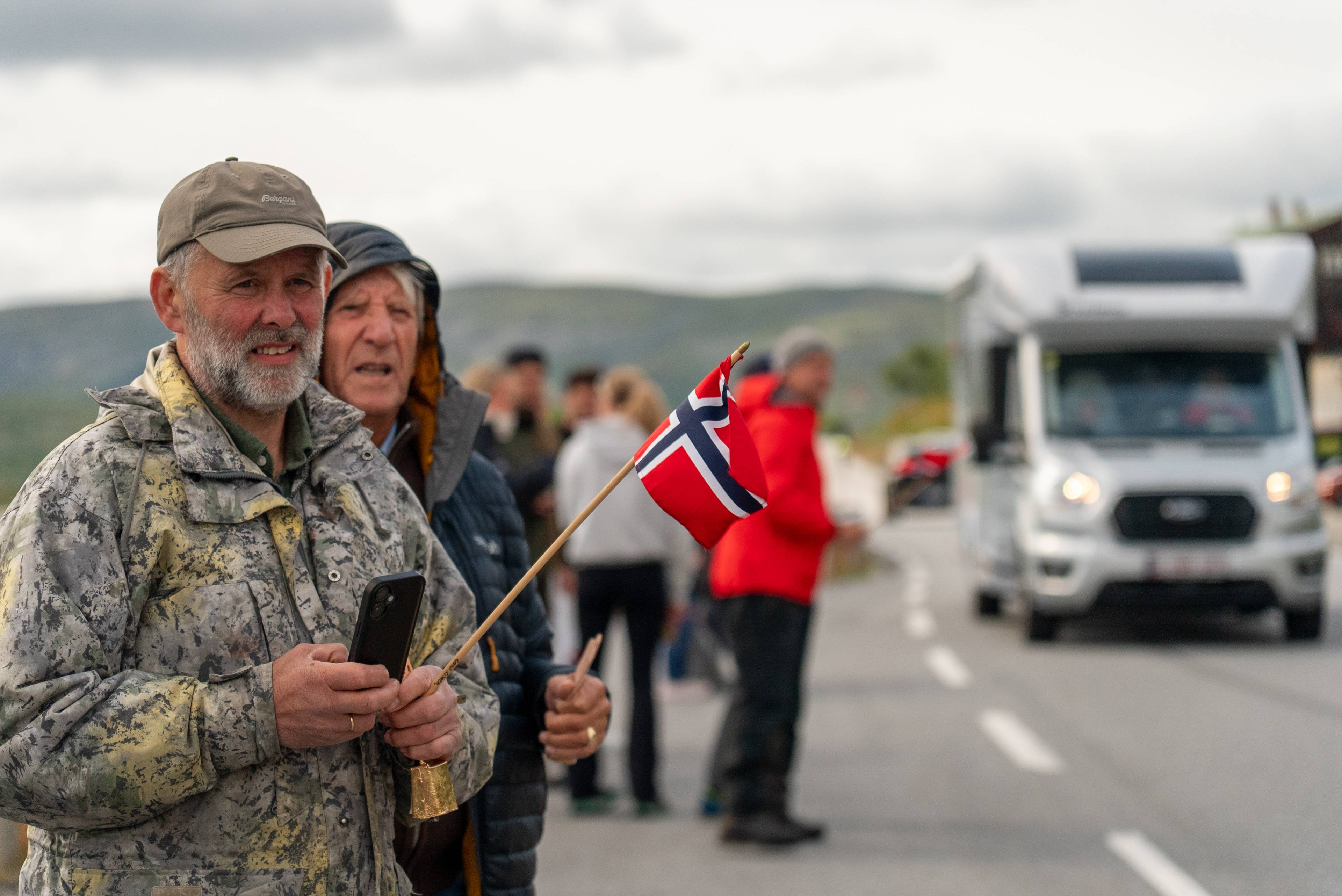Menn med et norsk flagg står langs en vei mens en bobil kjører forbi i bakgrunnen. (Bildebeskrivelsen er laget av en KI-tjeneste)