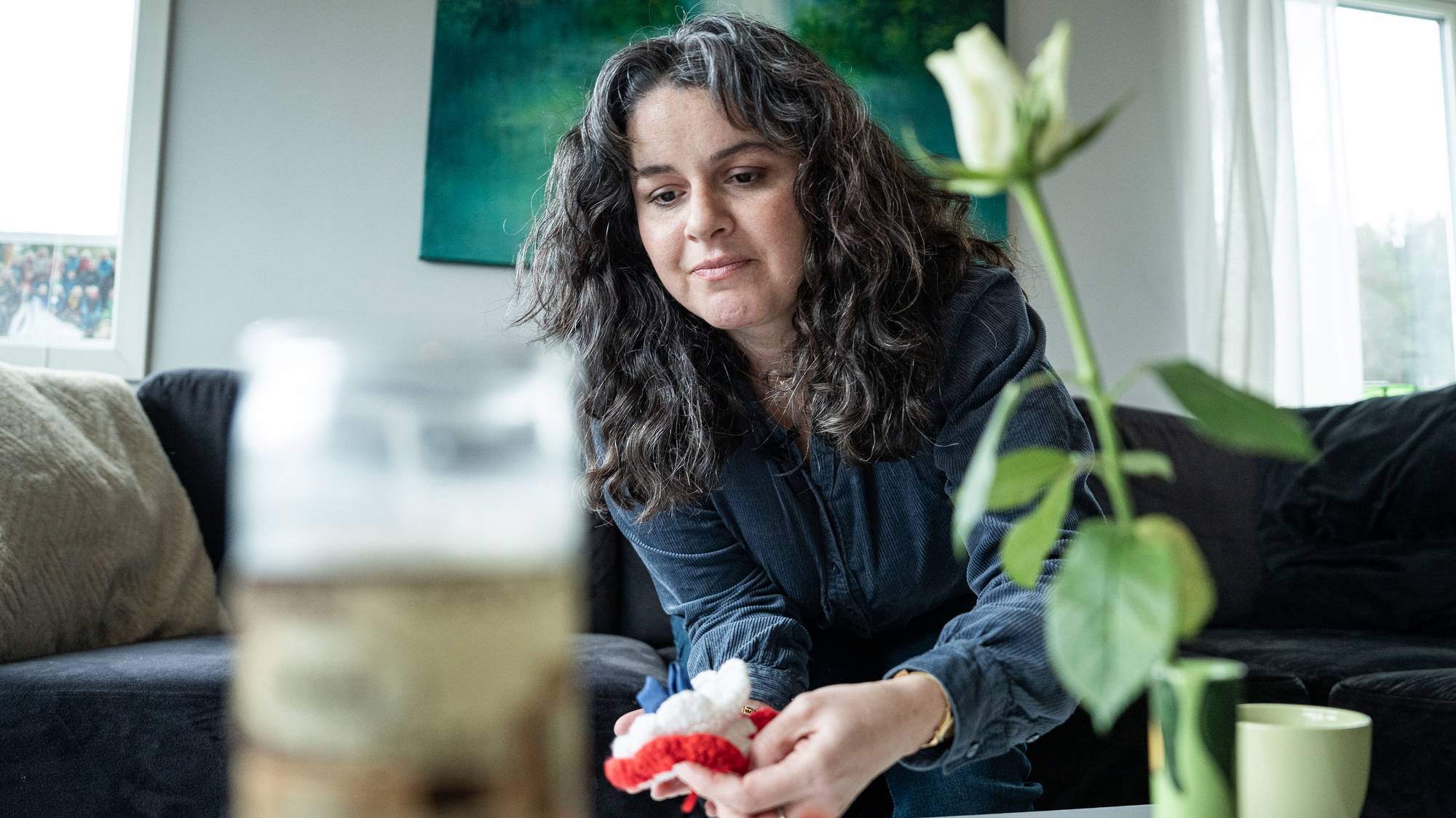 En kvinne sitter på en sofa og holder en liten, strikket gjenstand. Hun har mørkt, krøllete hår og ser ned på det hun holder. Foran henne står en hvit rose i en vase og en gul kopp. (Bildebeskrivelsen er laget av en KI-tjeneste)
