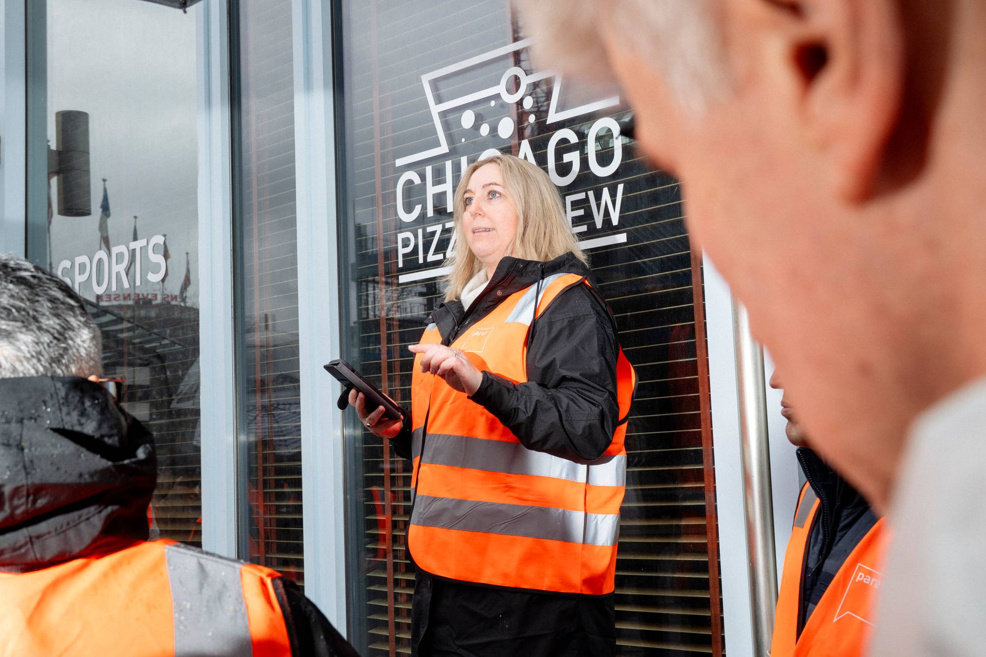 En kvinne i oransje vest holder en telefon mens hun snakker. Bak henne er det et vindu med en tekst som sier "CHICAGO PIZZA". Andre personer i oransje vester er i forgrunnen og til siden. (Bildebeskrivelsen er laget av en KI-tjeneste)
