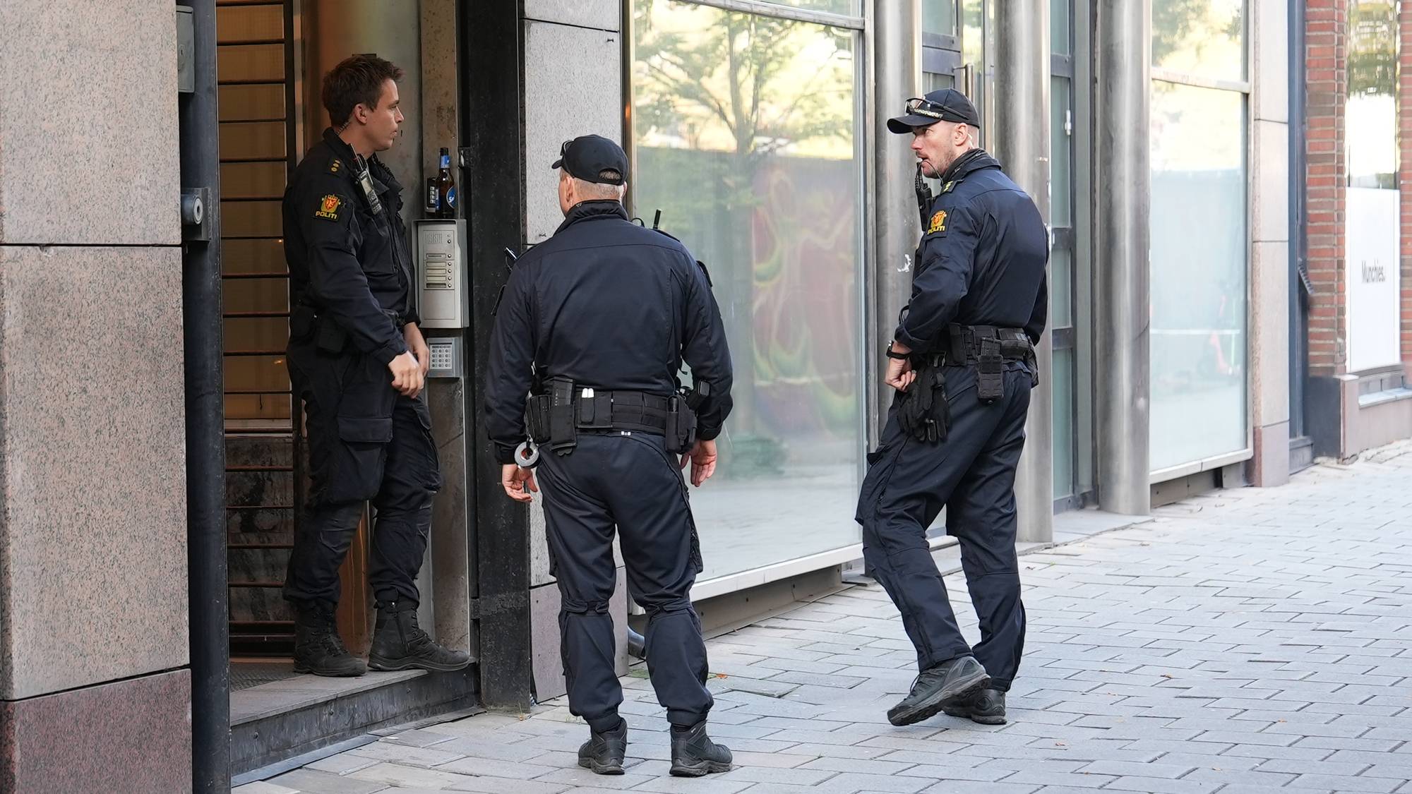 Tre politibetjenter i uniform står utenfor en bygningsinngang i Pilestredet. (Bildebeskrivelsen er laget av en KI-tjeneste)
