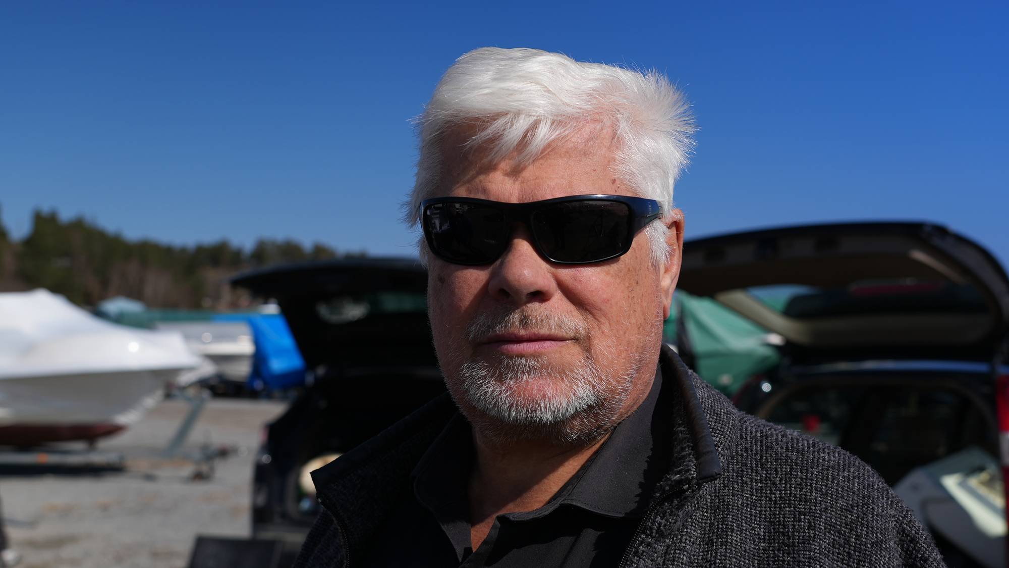 En mann med hvitt hår og solbriller ser inn i kamera. Han har på seg en mørk skjorte og en mørk jakke. Bak mannen sees et åpent bagasjerom på en bil. I bakgrunnen er det flere båter dekket til, trær og en klar blå himmel. (Bildebeskrivelsen er laget av en KI-tjeneste)