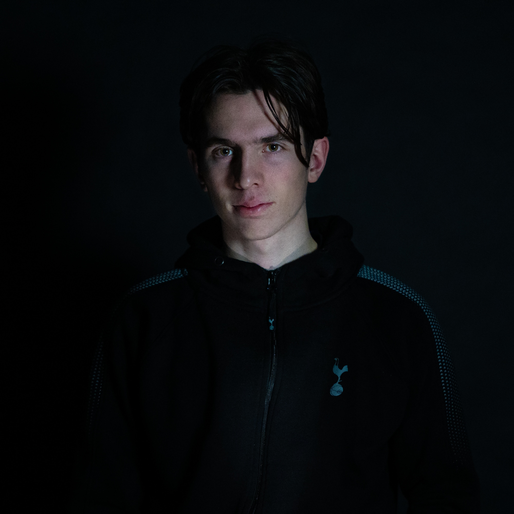Andreas (22) står i et helt mørkt rom. Kun ansiktet er lyst opp fra undersiden med et kaldt lyst. Han ser alvorlig inn i kameraet. Han har mørkt hår og lys hud. 