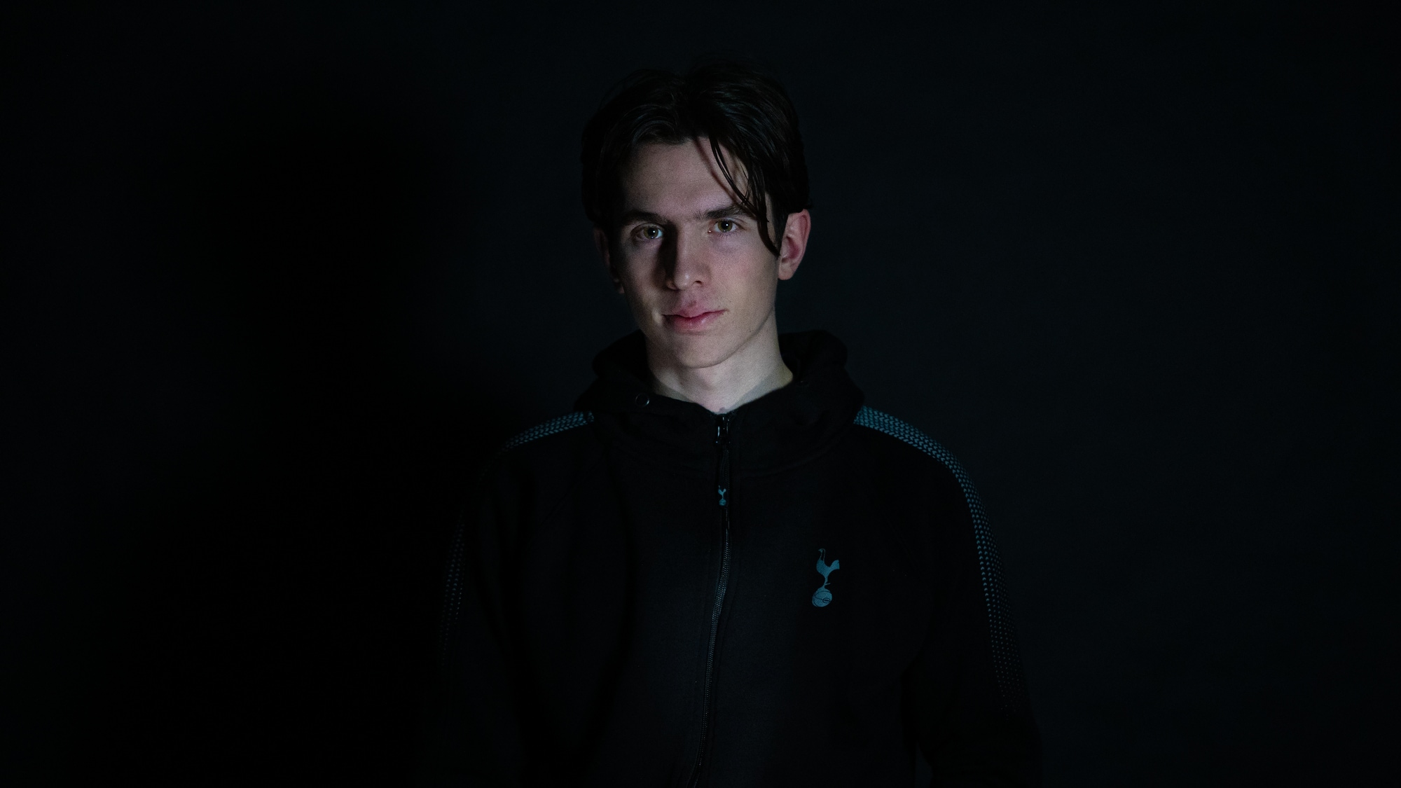 Andreas (22) står i et helt mørkt rom. Kun ansiktet er lyst opp fra undersiden med et kaldt lyst. Han ser alvorlig inn i kameraet. Han har mørkt hår og lys hud. 