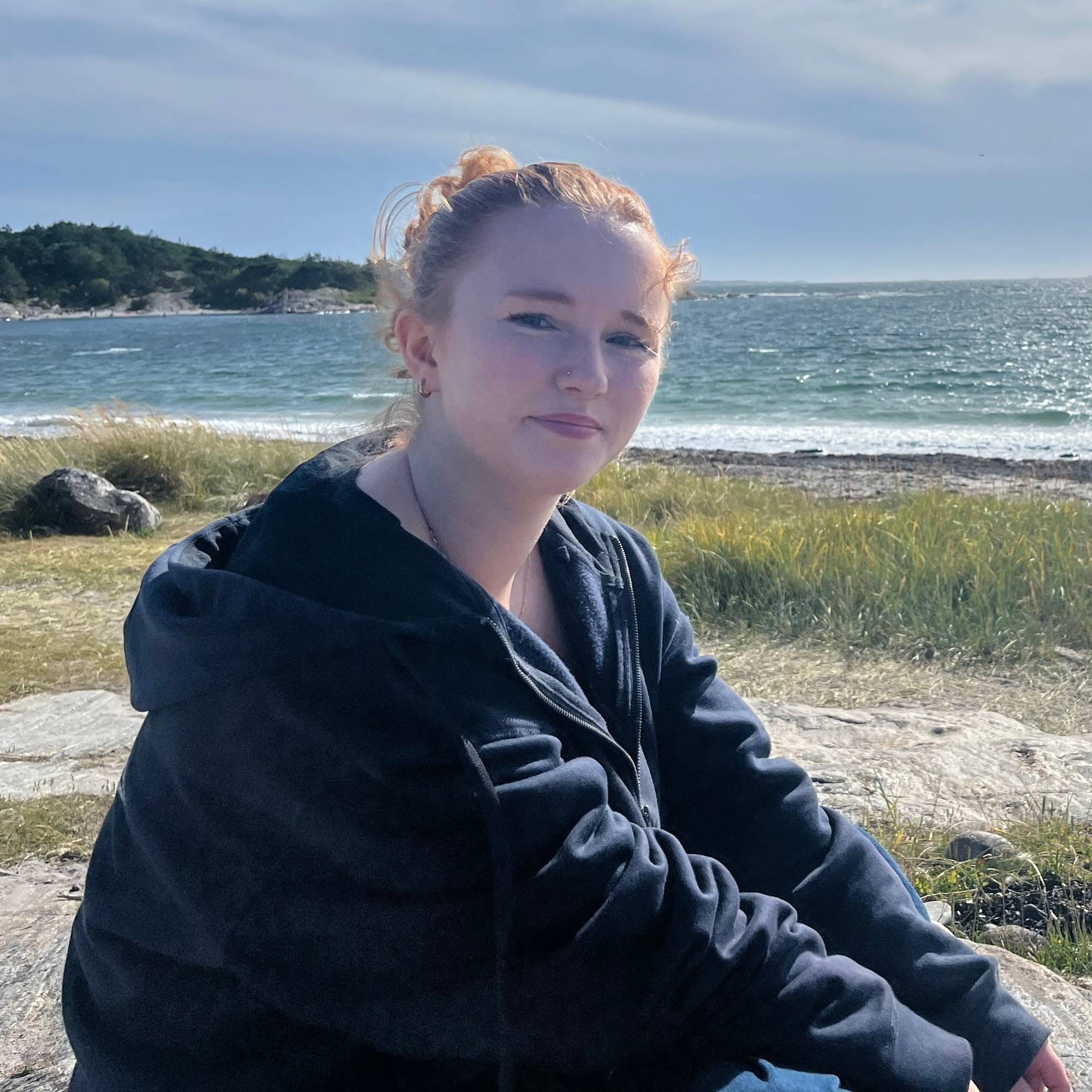 Kvinne med rødt hår sitter på et svaberg ved en strand med havet i bakgrunnen. (Bildebeskrivelsen er laget av en KI-tjeneste)