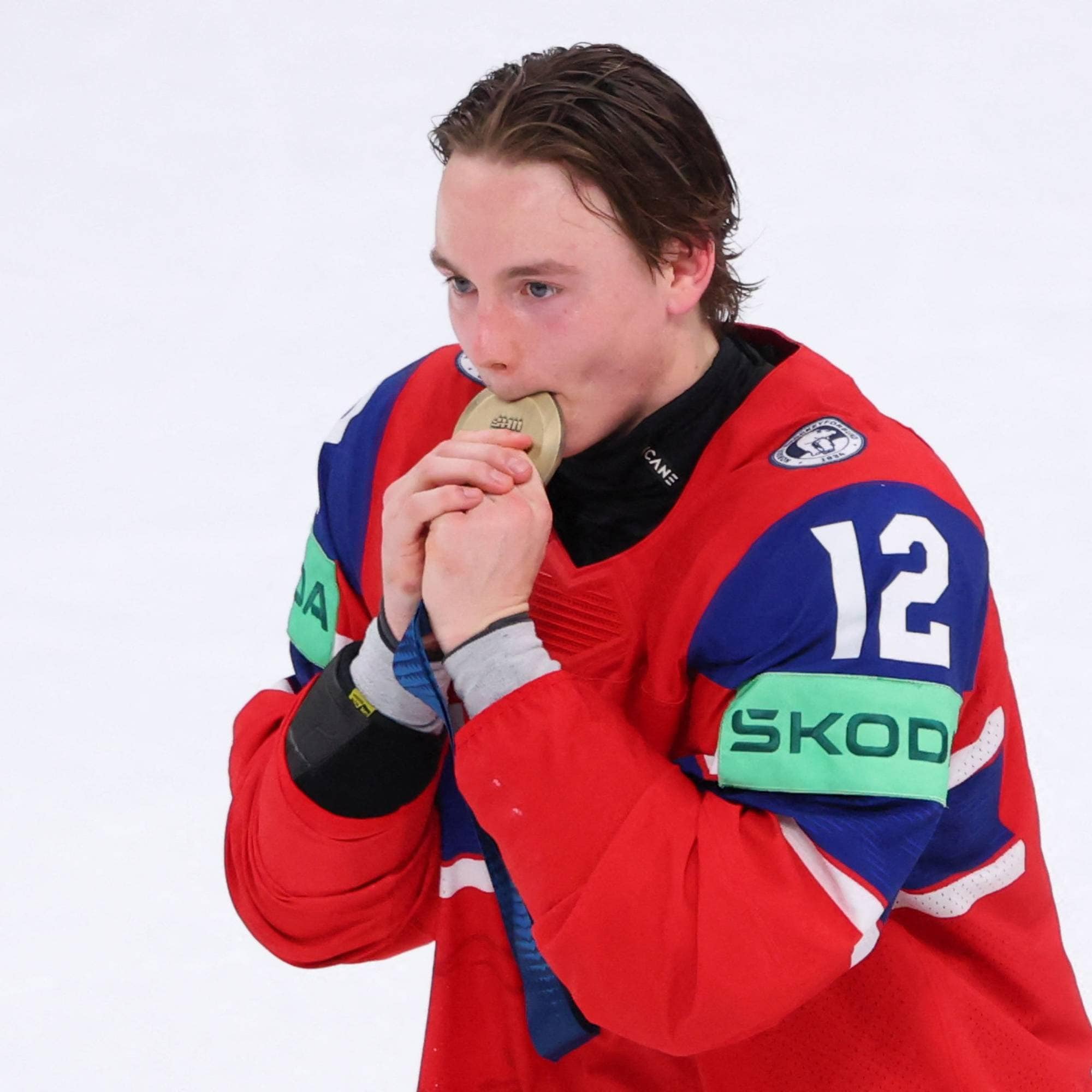 En ishockeyspiller i en rød drakt med nummer 12 biter på en gullmedalje. Spilleren har hendene rundt medaljen. Det er et hvitt bakteppe. (Bildebeskrivelsen er laget av en KI-tjeneste)