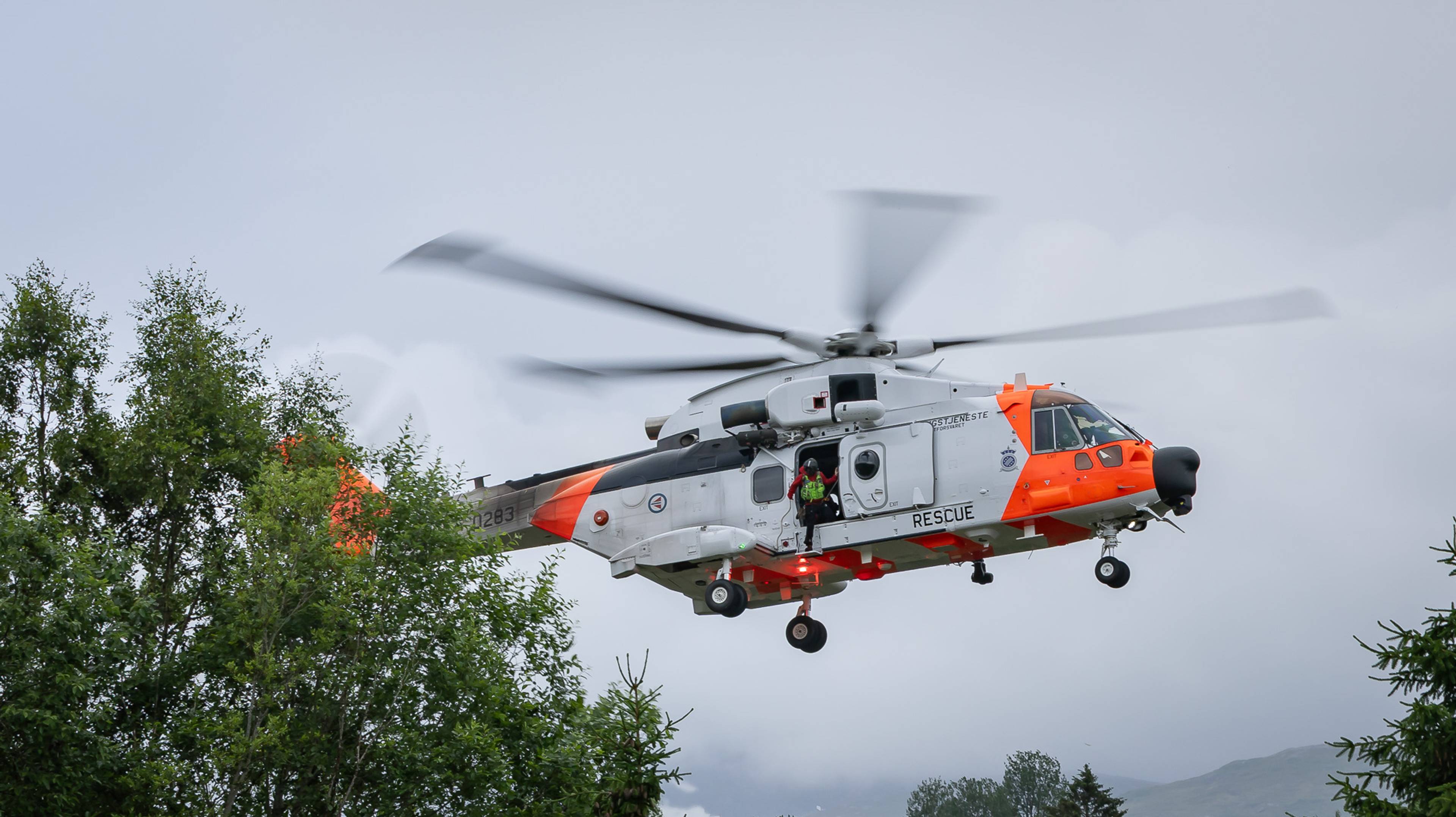 Et AW101 SAR Queen redningshelikopter er i ferd med å lande. Vi ser en person med hjelm og kommunikasjonsutstyr i døråpningen. Vedkommende kikker ned. I forkant av bildene ser vi toppen av noen trær, og i bakgrunnen kan vi se deler av et fjell og antydninger til lavt skydekke