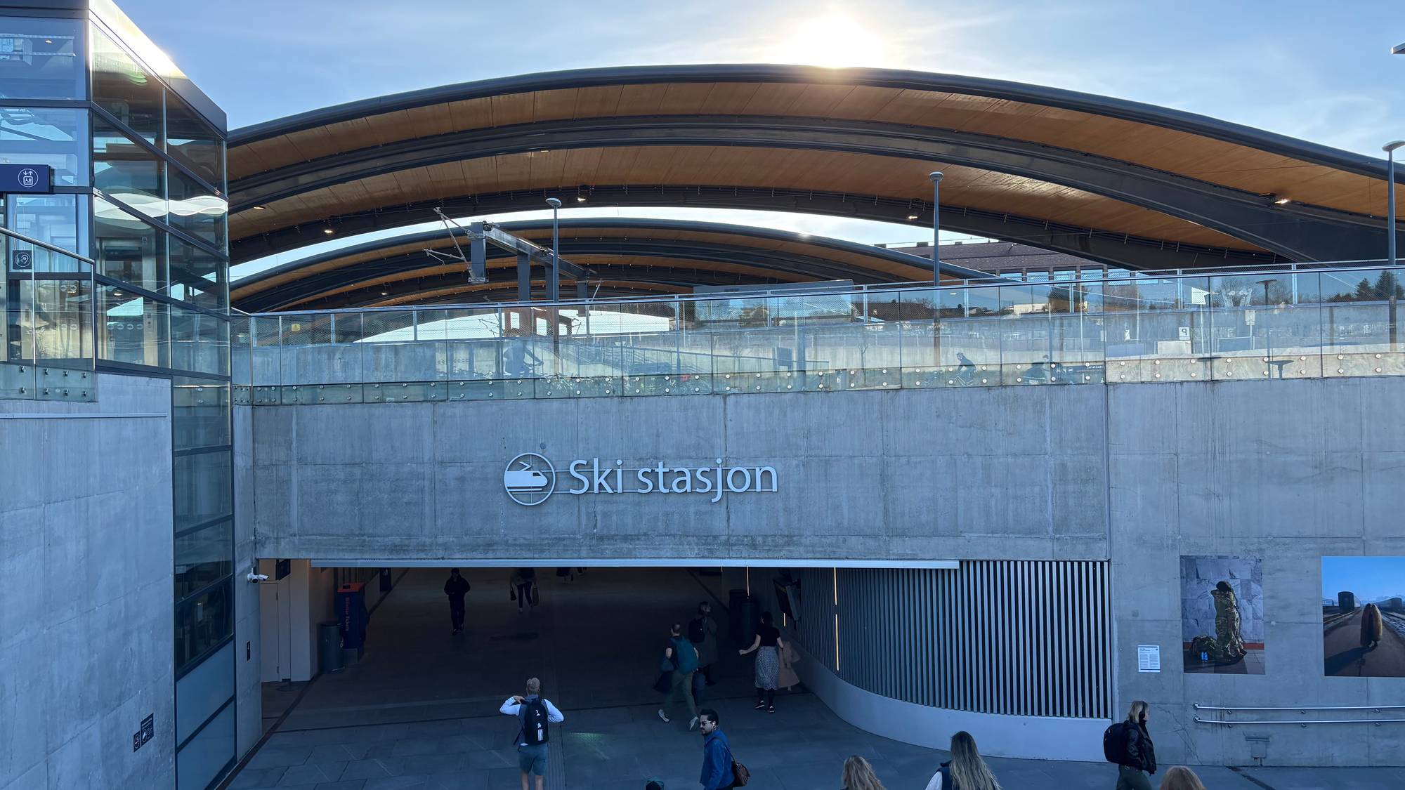 Folk går inn i en bygning merket Ski stasjon. Bygningen har en moderne arkitektur med betong og glass. Solen skinner fra en klar himmel over den bøyde takkonstruksjonen. (Bildebeskrivelsen er laget av en KI-tjeneste)
