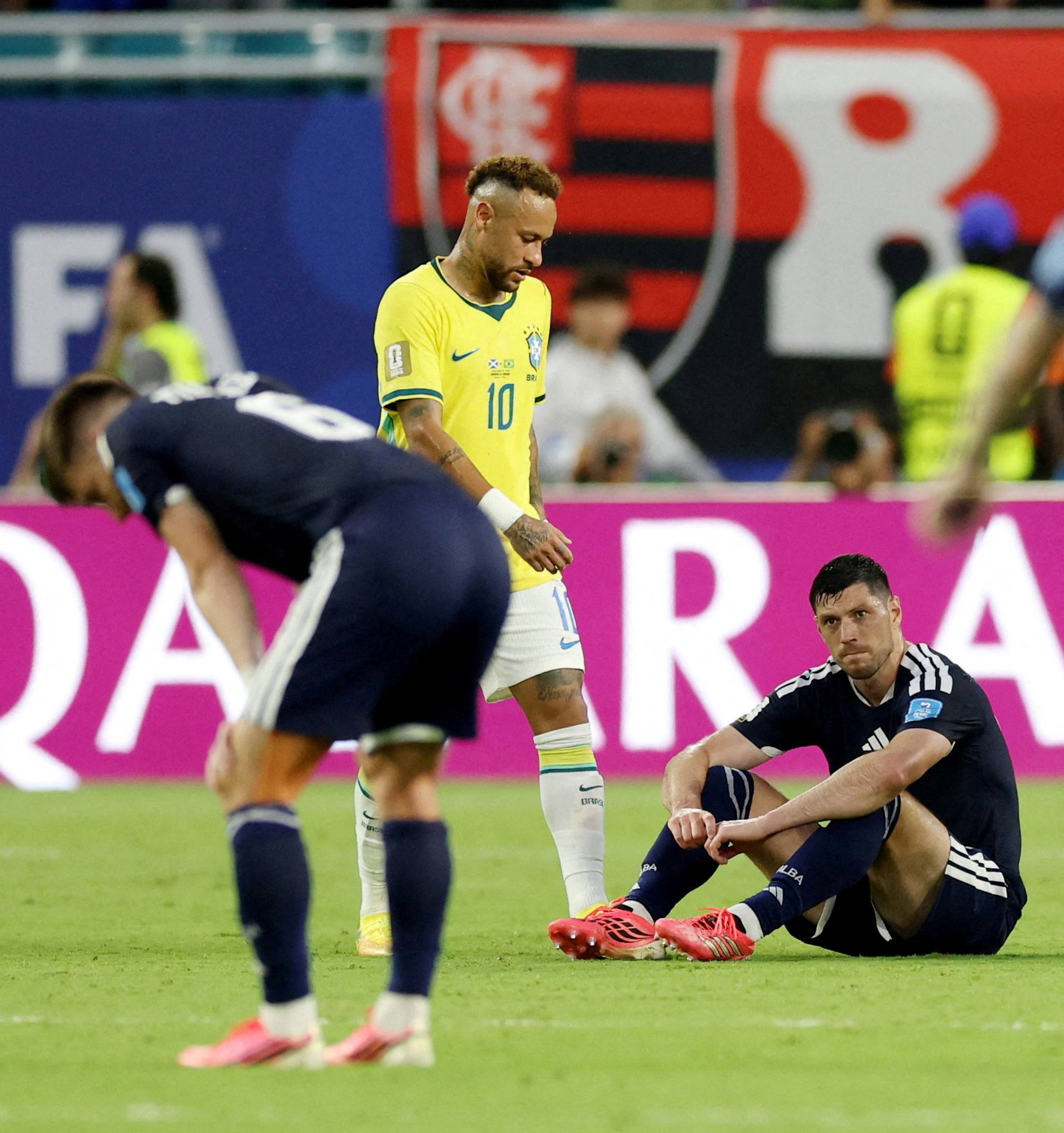 Fotballspilleren Neymar i gul drakt med nummer 10 står på en gressbane. To spillere i mørke drakter sitter eller bøyer seg ned. Det er en banner med "QATAR" i bakgrunnen. (Bildebeskrivelsen er laget av en KI-tjeneste)
