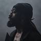 Sony Music Norway / Sony Music Norway En mann står i profil mot en uklar, dyster bakgrunn. Han har på seg en mørk jakke og en hodeplagg. Huden er synlig, sammen med en kort, velstelt skjeggvekst. Belysningen gir et dramatisk uttrykk i bildet. (Bildebeskrivelsen er laget av en KI-tjeneste)