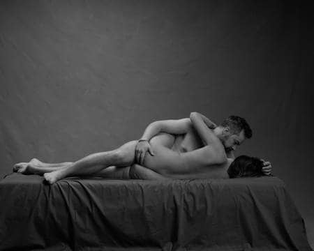 En naken mann med mørkt hår og skjegg ligger på siden på senga med høyrebenet mellom bena på en naken dame som holder rundt ham