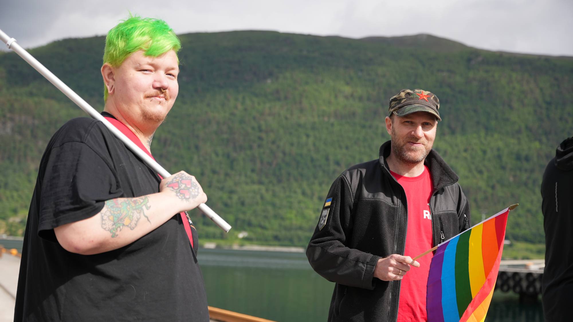 To personer står ved en brygge. Den ene har grønt hår og holder en hvit stang. Den andre har på seg caps og holder et regnbueflagg. I bakgrunnen er en fjellside med grønn vegetasjon. (Bildebeskrivelsen er laget av en KI-tjeneste)