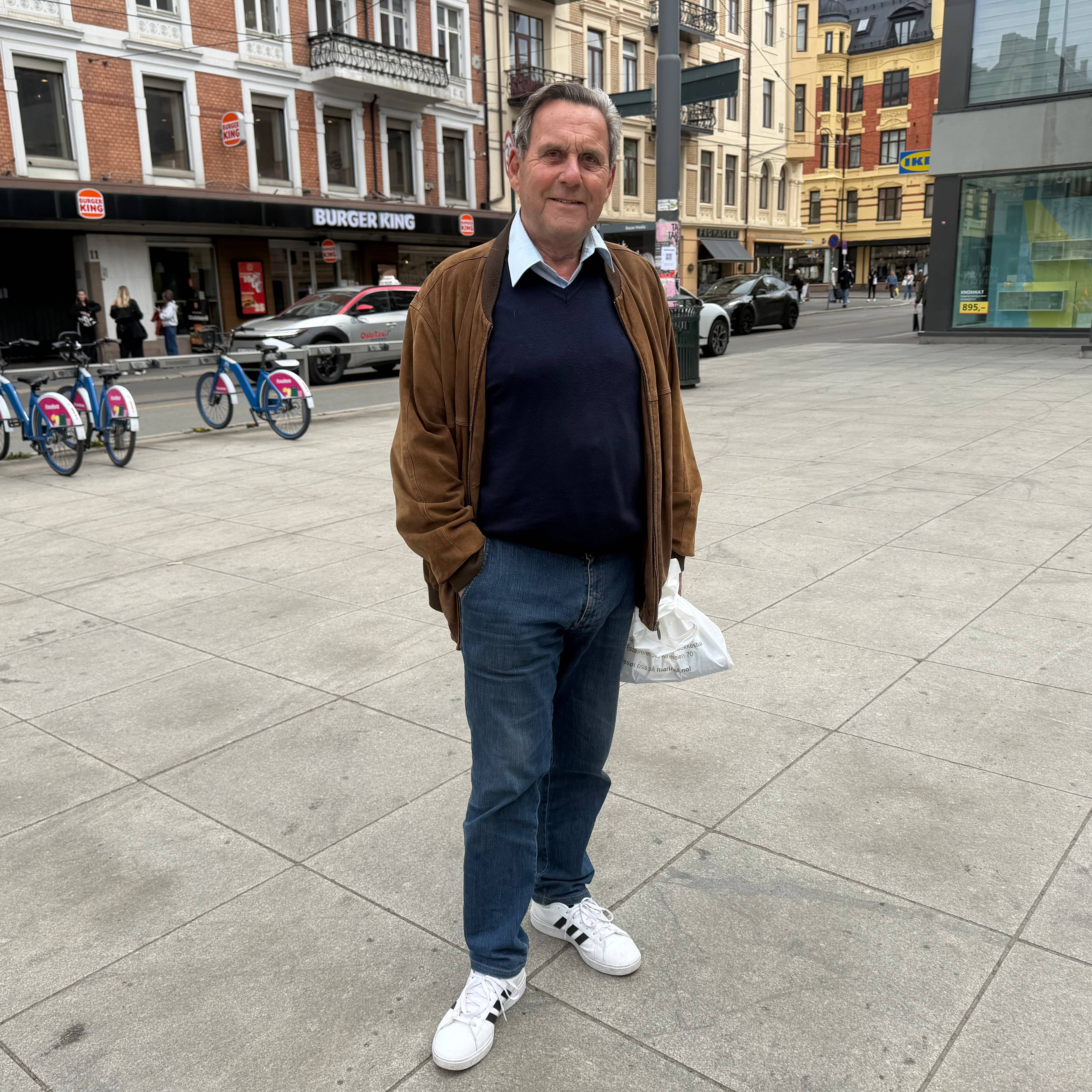 En mann står i en gate med sykler og bygninger bak seg. Han har på seg en brun jakke, mørk genser og blå jeans. I bakgrunnen sees en Burger King og en IKEA-butikk. (Bildebeskrivelsen er laget av en KI-tjeneste)