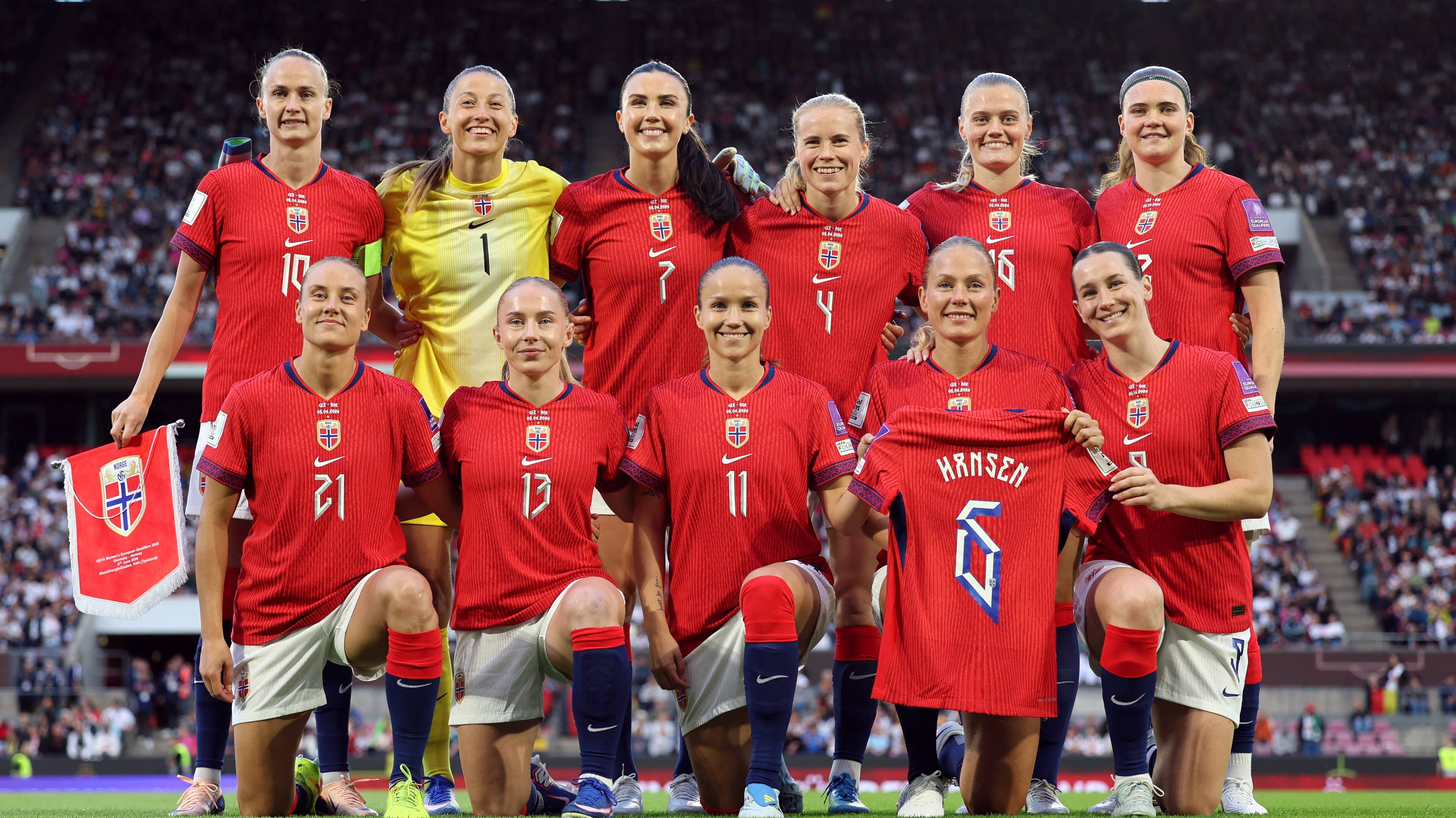 Norges kvinnelandslag i fotball poserer foran et publikum. Spillerne har på seg røde draktsett og hvite shorts. En spiller holder opp en trøye med navnet "Hansen" og nummer 6. (Bildebeskrivelsen er laget av en KI-tjeneste)