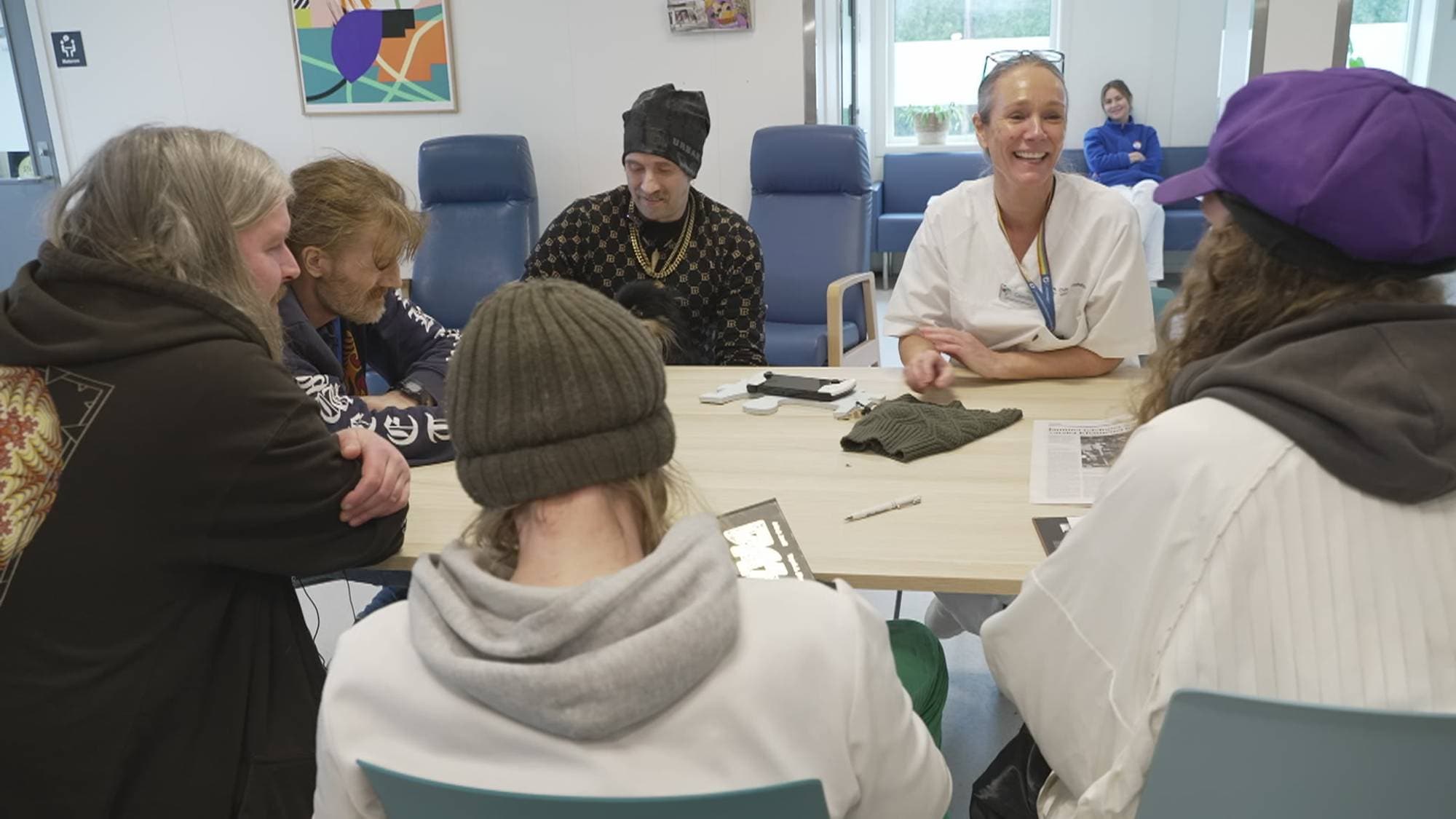 En gruppe mennesker sitter rundt et bord. En kvinne i helsepersonelluniform ler mens hun ser på en av de andre. En mann med gullkjede sitter ved siden av henne. (Bildebeskrivelsen er laget av en KI-tjeneste)