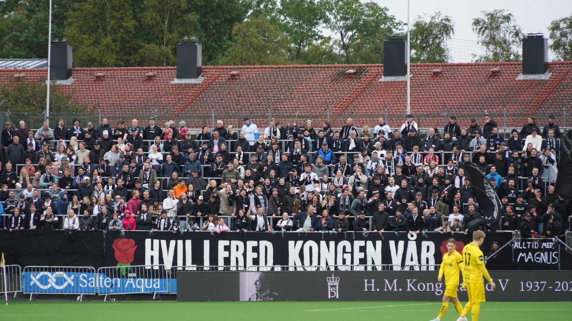 Supportere på en fotballtribune holder opp et banner til minne om kong Harald V. (Bildebeskrivelsen er laget av en KI-tjeneste)