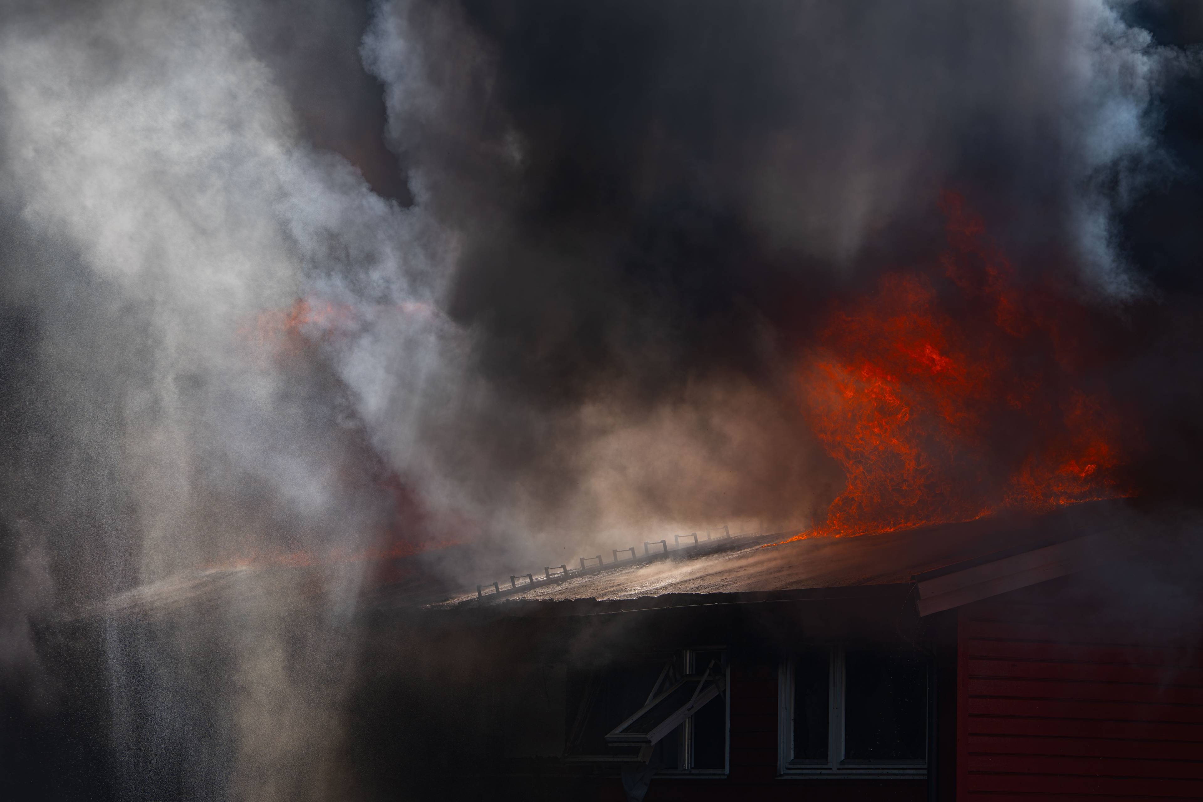 En bygning står i flammer med store mengder røyk. Rødt og oransje lys fra flammene lyser opp taket. Vann sprøytes mot røyken fra venstre. (Bildebeskrivelsen er laget av en KI-tjeneste)