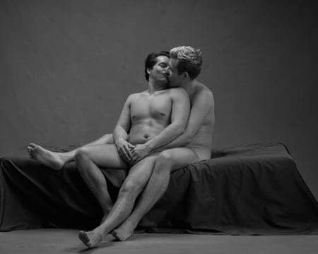 En naken mann med mørkt hår sitter på fanget til en annen naken mann som holder rundt ham. De kysser og tar på hverandre.