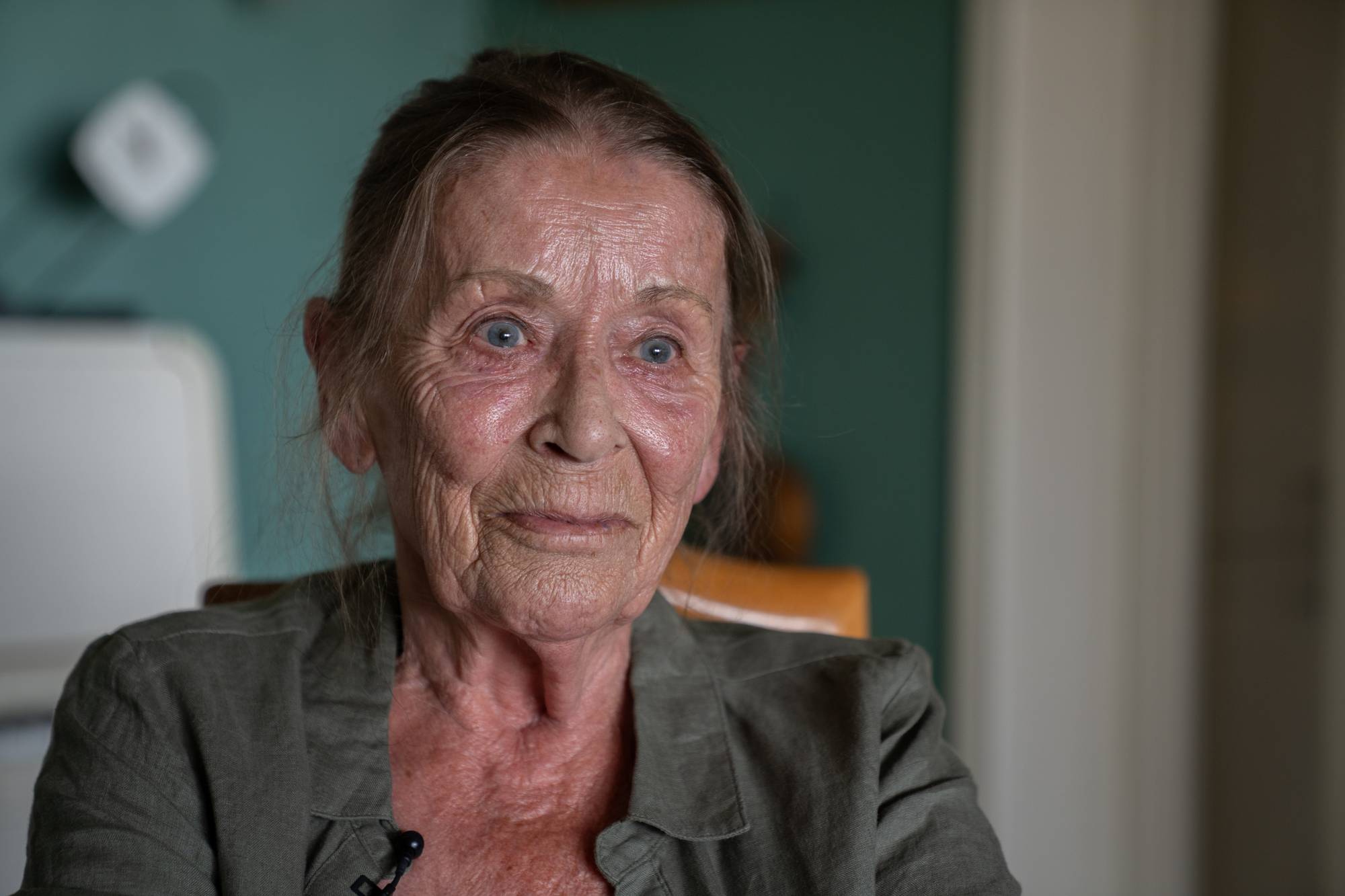 Portrett av en solbrun, eldre kvinne. Hun sitter inne, har oppsatt, mørkt hår og lys, grønn skjorte.
