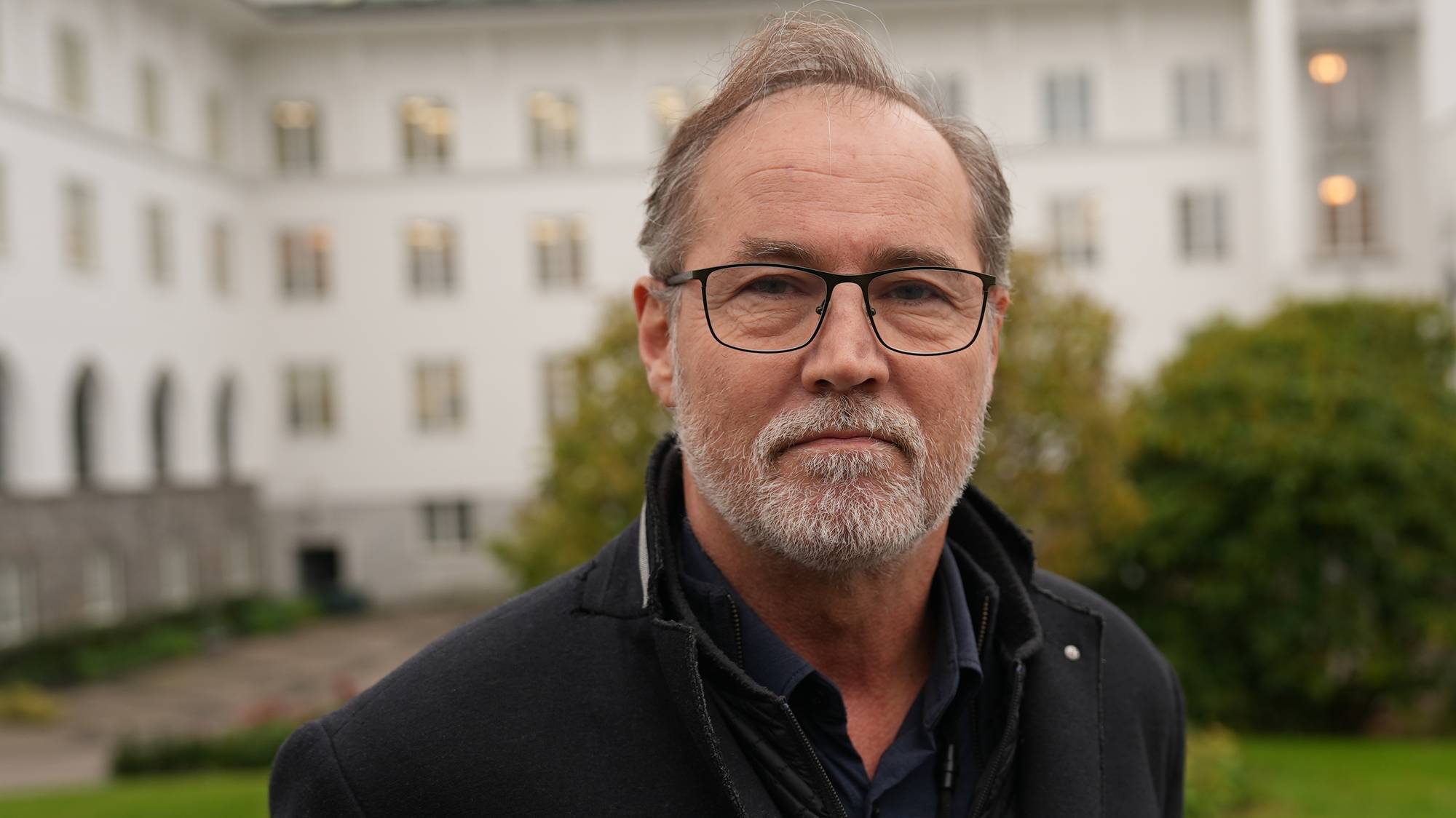 Portrett av en mann med briller og skjegg foran en hvit bygning. (Bildebeskrivelsen er laget av en KI-tjeneste)