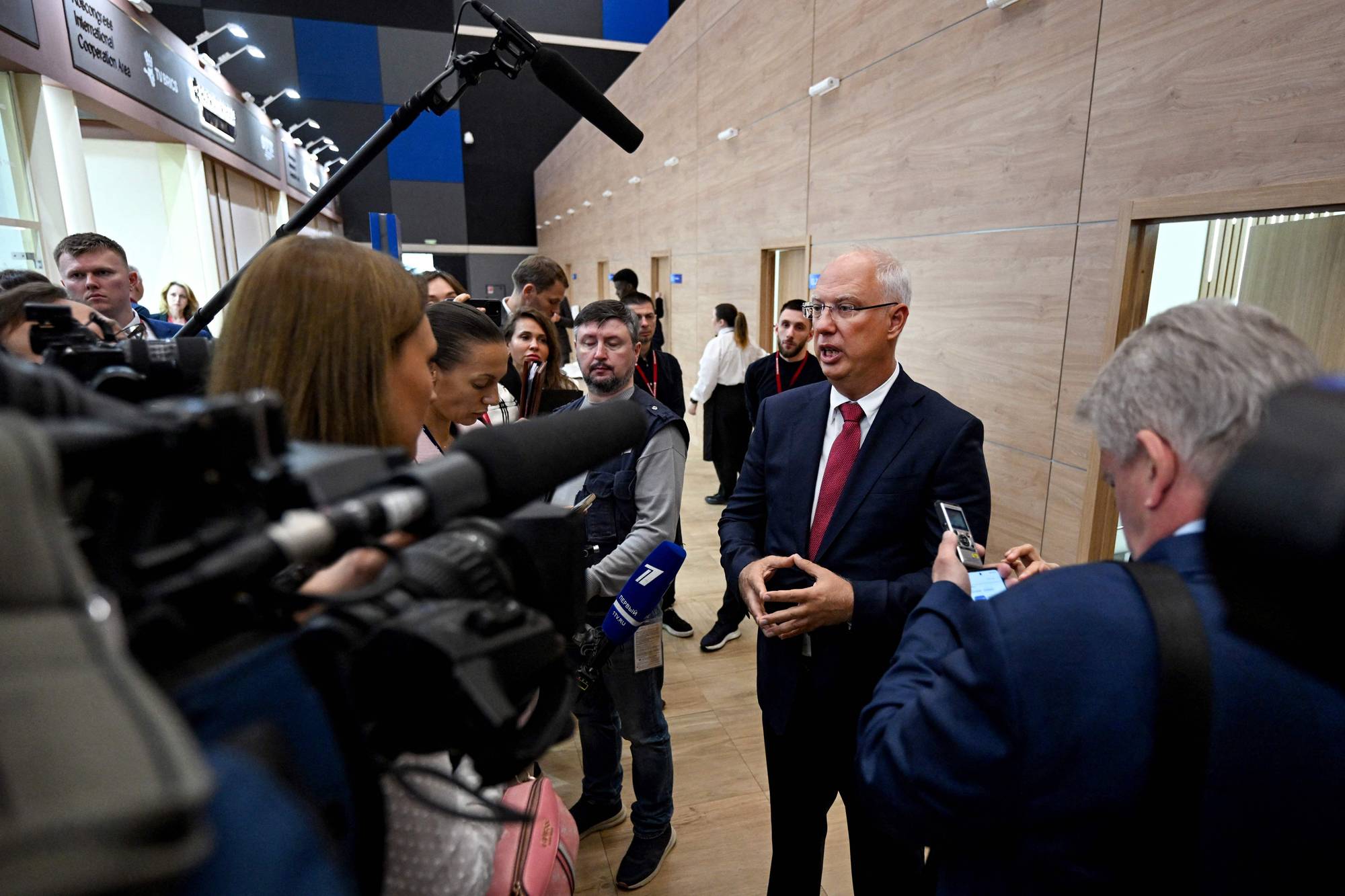 En mann i dress og slips snakker med journalister. Flere kameraer og mikrofoner er rettet mot ham. Omgivelsene er en moderne bygning med trepanel på veggene. (Bildebeskrivelsen er laget av en KI-tjeneste)